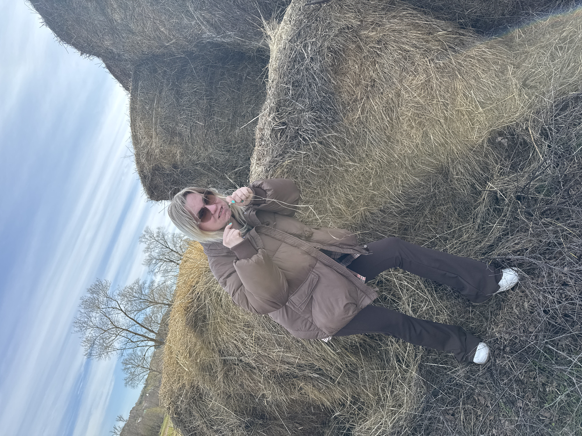 A woman is reclining against a stack of hay bales. She is wearing sunglasses, a brown puffer jacket, brown pants, and white sneakers. She is holding a piece of straw in her hands. The location appears to be a rural area near the city of Уральское, Russia, with a stack of hay bales, dry grass, and bare trees in the background under a partly cloudy sky.