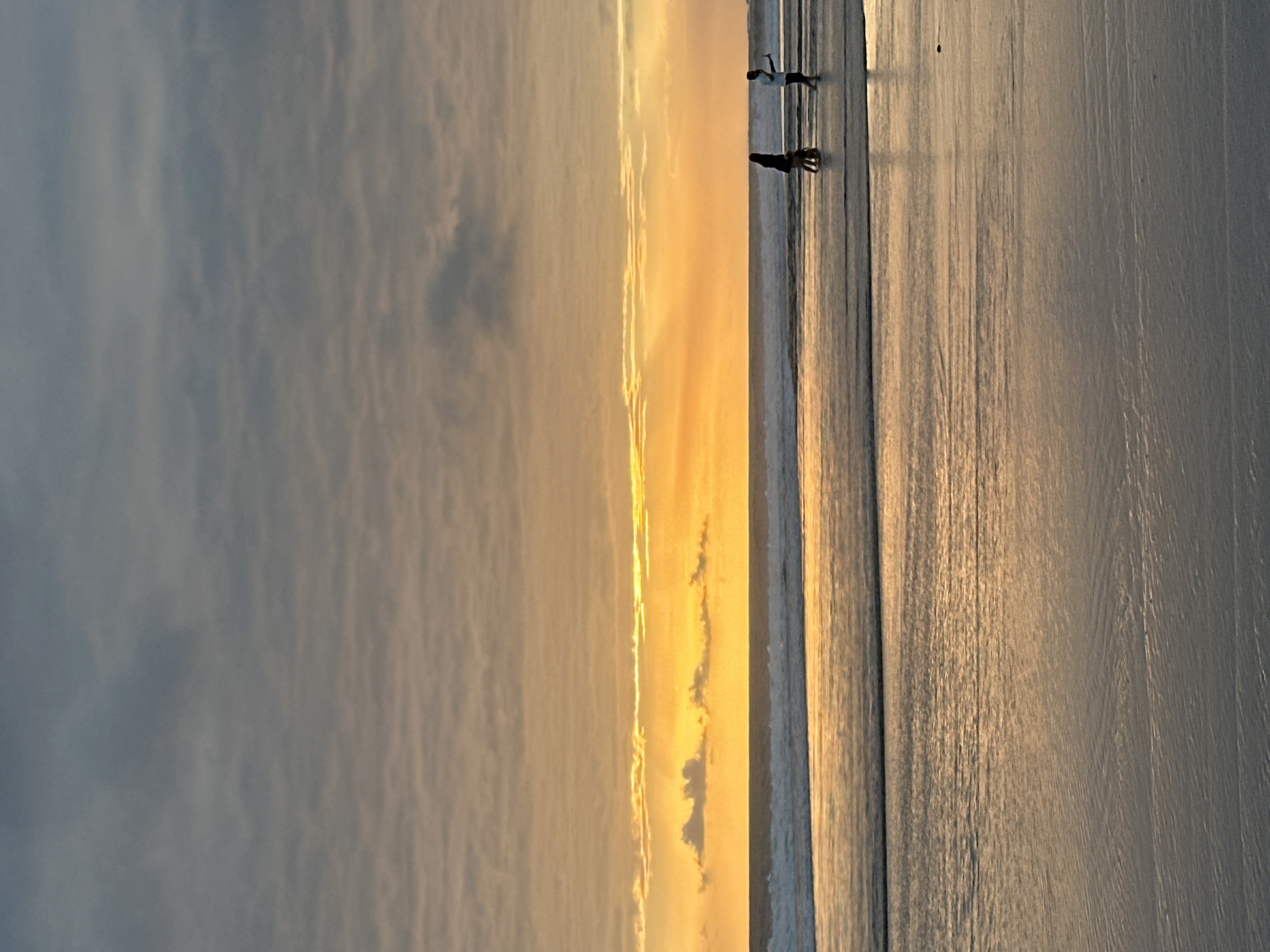 The image depicts a serene beach scene at sunset in North Kuta, Indonesia. The sky is filled with a dramatic gradient of colors, transitioning from a dark, cloudy gray at the top to a vibrant, glowing orange near the horizon. Wispy clouds are scattered across the sky, some catching the golden light.
In the mid-ground, the ocean meets the shore, with gentle waves rolling onto the wet sand. The water reflects the warm hues of the sunset, creating a shimmering effect.
On the beach, two figures are visible. To the left, a person in darker clothing appears to be walking with a smaller figure, possibly a child or a dog, next to them. To the right, a person in lighter clothing is standing, seemingly engaged with a device in their hand, perhaps taking a photo.
The foreground consists of the wet sand of the beach, marked with shallow ripples left by the receding tide. The overall atmosphere is peaceful and tranquil, characteristic of a sunset on a tropical beach.