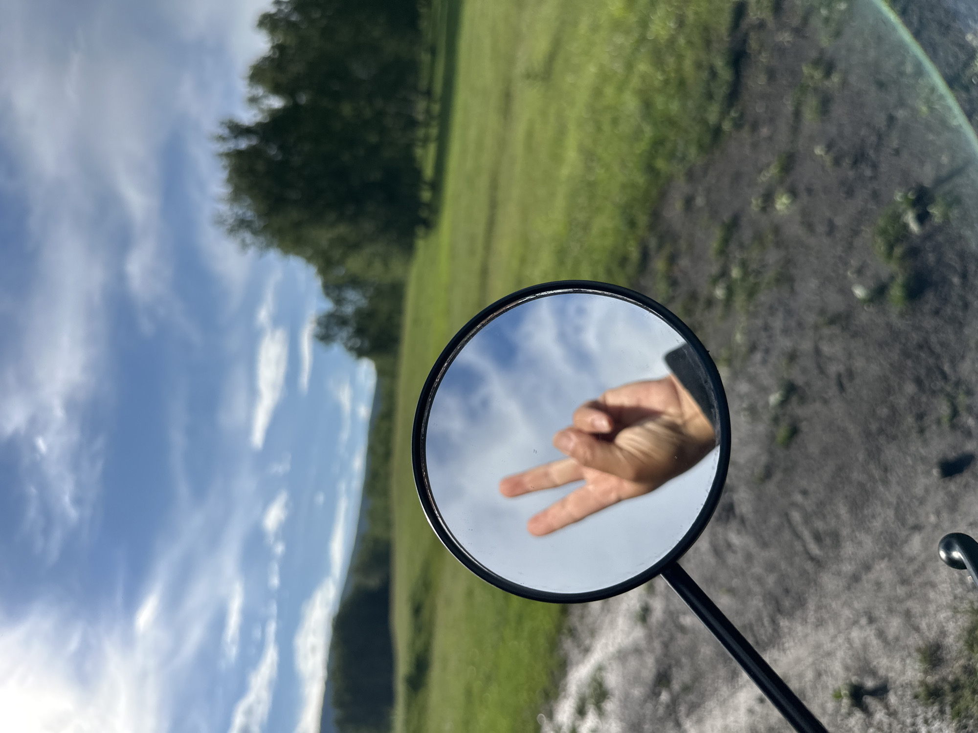 An extreme close-up shot shows a reflection of a hand with two fingers raised in a peace sign in a motorcycle mirror. The mirror is round and black, attached to a black stem. The background is a blurred landscape of green fields, trees, and a blue sky with white clouds. The ground in the foreground appears to be dirt and gravel.