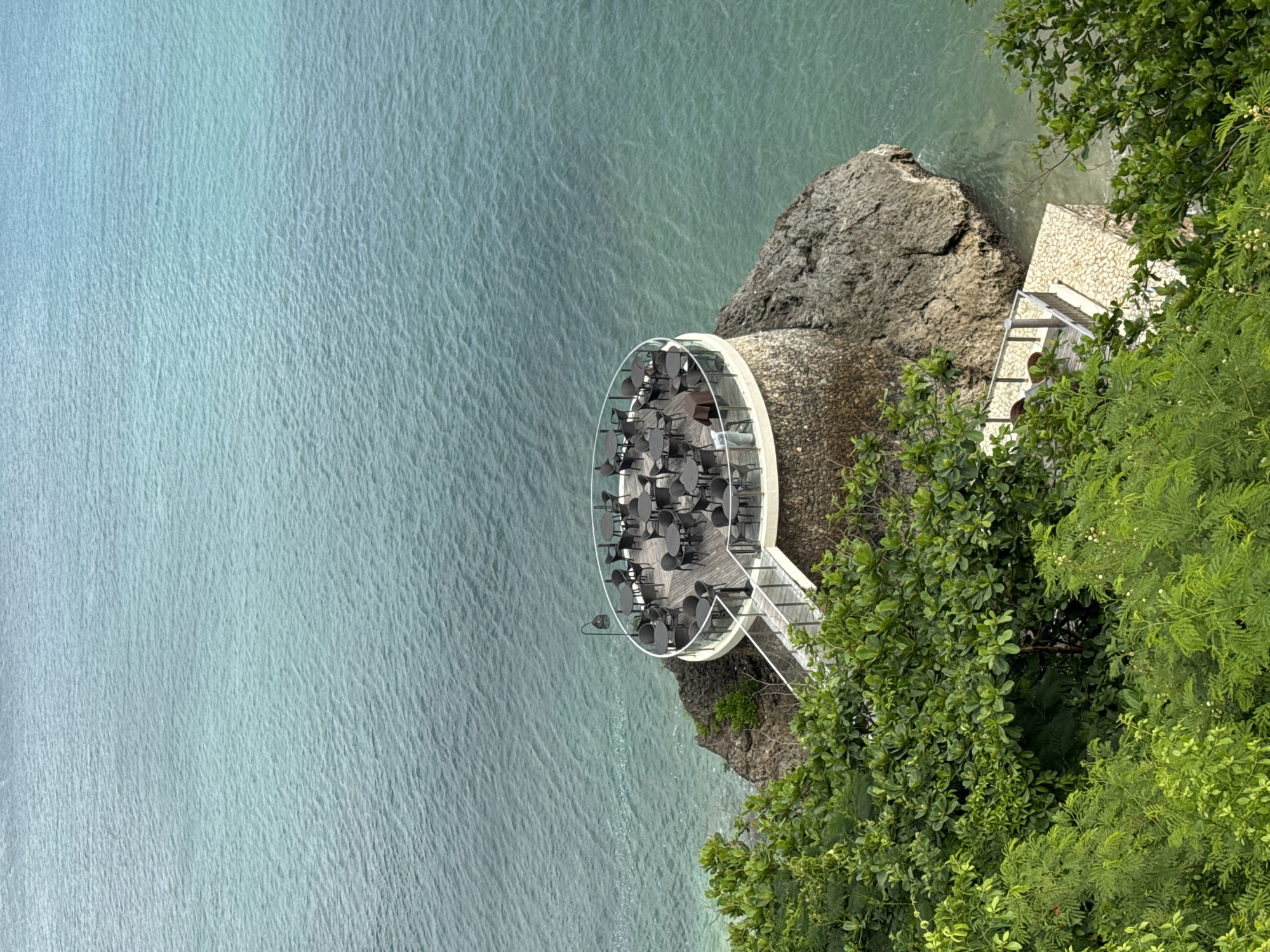An aerial shot captures the Rock Bar at Ayana Resort in Kuta Selatan, Indonesia. The circular bar, furnished with tables and chairs, is positioned on a rocky cliff edge overlooking the turquoise ocean. Lush green foliage surrounds the bar area on the right side of the image.