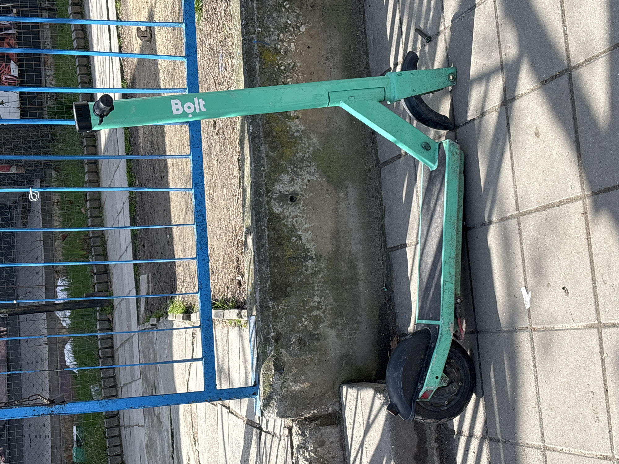 A Bolt electric scooter lies on a paved sidewalk in Varna, Bulgaria. The scooter is light green with the word "Bolt" printed on the frame. It rests against a blue metal fence. The sidewalk appears to be made of gray paving stones, and there is a narrow strip of dirt and concrete separating the sidewalk from the fence. A small bolt lies on the sidewalk near the front wheel. The scene is brightly lit, suggesting it is daytime.
