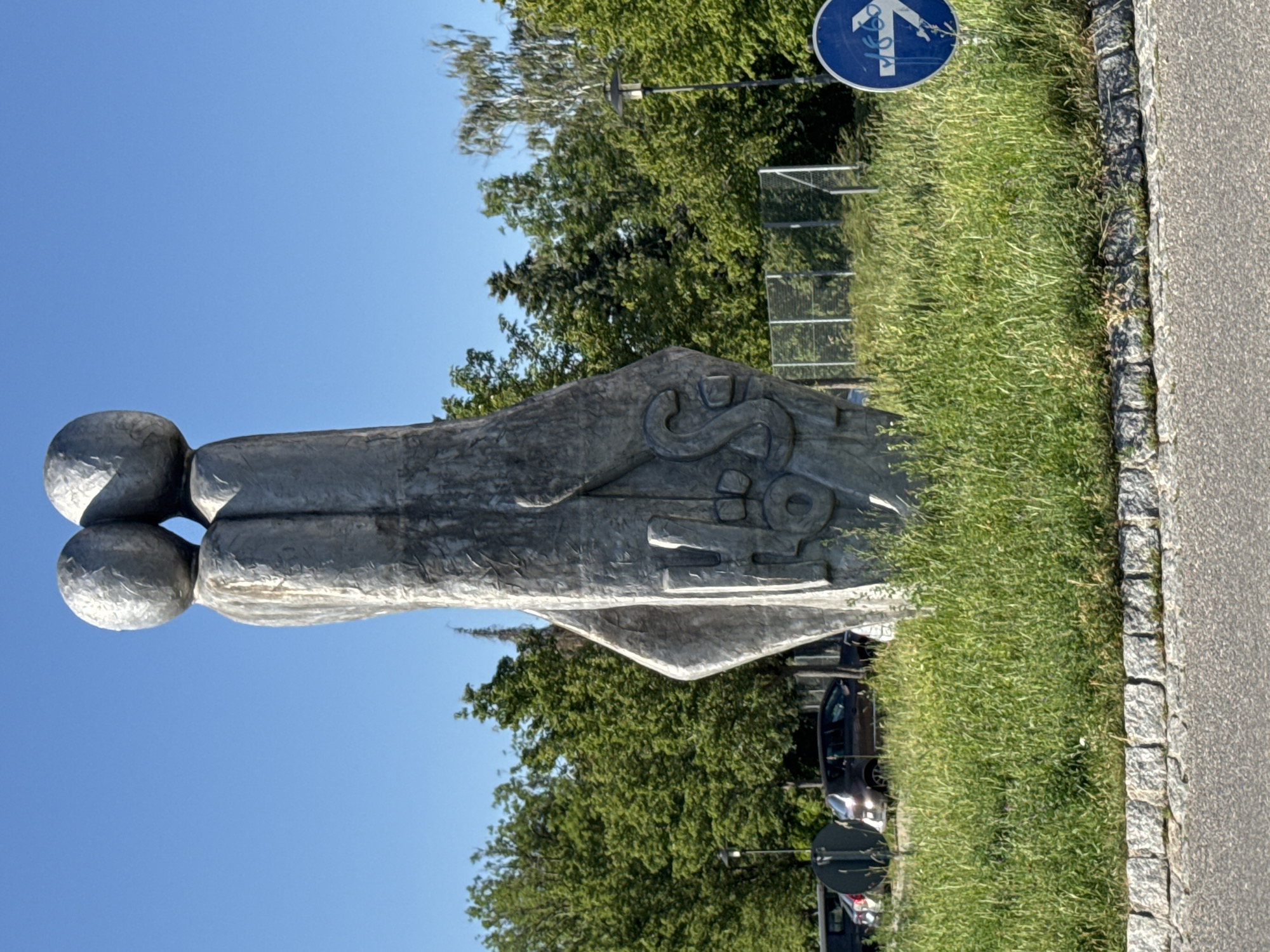 This media file depicts a large, abstract outdoor sculpture situated beside a paved road and a grassy area. The sculpture appears to be made of a rough, grey material, possibly concrete or stone. It has a smooth, rounded top section resembling two spheres joined together, and a more angular, textured lower section with carved-out shapes that vaguely resemble stylized letters or symbols.
Behind the sculpture, lush green trees form a backdrop against a clear blue sky, suggesting a daytime scene. A paved pathway or road runs along the right side of the sculpture, bordered by rough-hewn stones. To the left of the road and in front of the trees, a patch of vibrant green grass is visible.
In the upper right corner, a blue circular sign with a white bicycle symbol is mounted on a pole, indicating a bicycle route. A metal fence or barrier is also visible behind the grass and trees. Several cars are parked or driving in the background, partially obscured by the trees, suggesting an urban or suburban setting. The lighting indicates a sunny day, possibly late morning or afternoon, with no clouds visible. The overall impression is a public space featuring a piece of modern art within a natural setting. The context provided places this scene in Höhenkirchen-Siegertsbrunn, Germany.