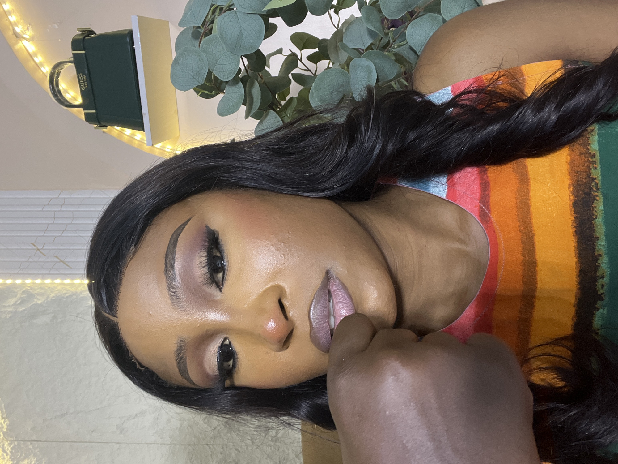 A close-up, eye-level selfie shows a woman with dark hair, styled in loose waves, wearing a brightly colored, striped tank top. Her face is angled towards the camera, with a visible portion of her left hand resting on her chin, obscuring her mouth. She has on dramatic eye makeup, including winged eyeliner and false eyelashes, and a shimmering, pale pink lipstick.
In the background, a decorative shelf holds a dark green Guess handbag. To the right of the handbag, a sprig of artificial eucalyptus branches is visible. The wall behind the woman appears to be a textured, light-colored surface, with a string of warm-toned fairy lights draped above the shelf. The scene is illuminated by soft lighting, creating a warm and inviting atmosphere. The photograph is taken indoors, with no specific landmarks indicating it is in Abuja, Nigeria.