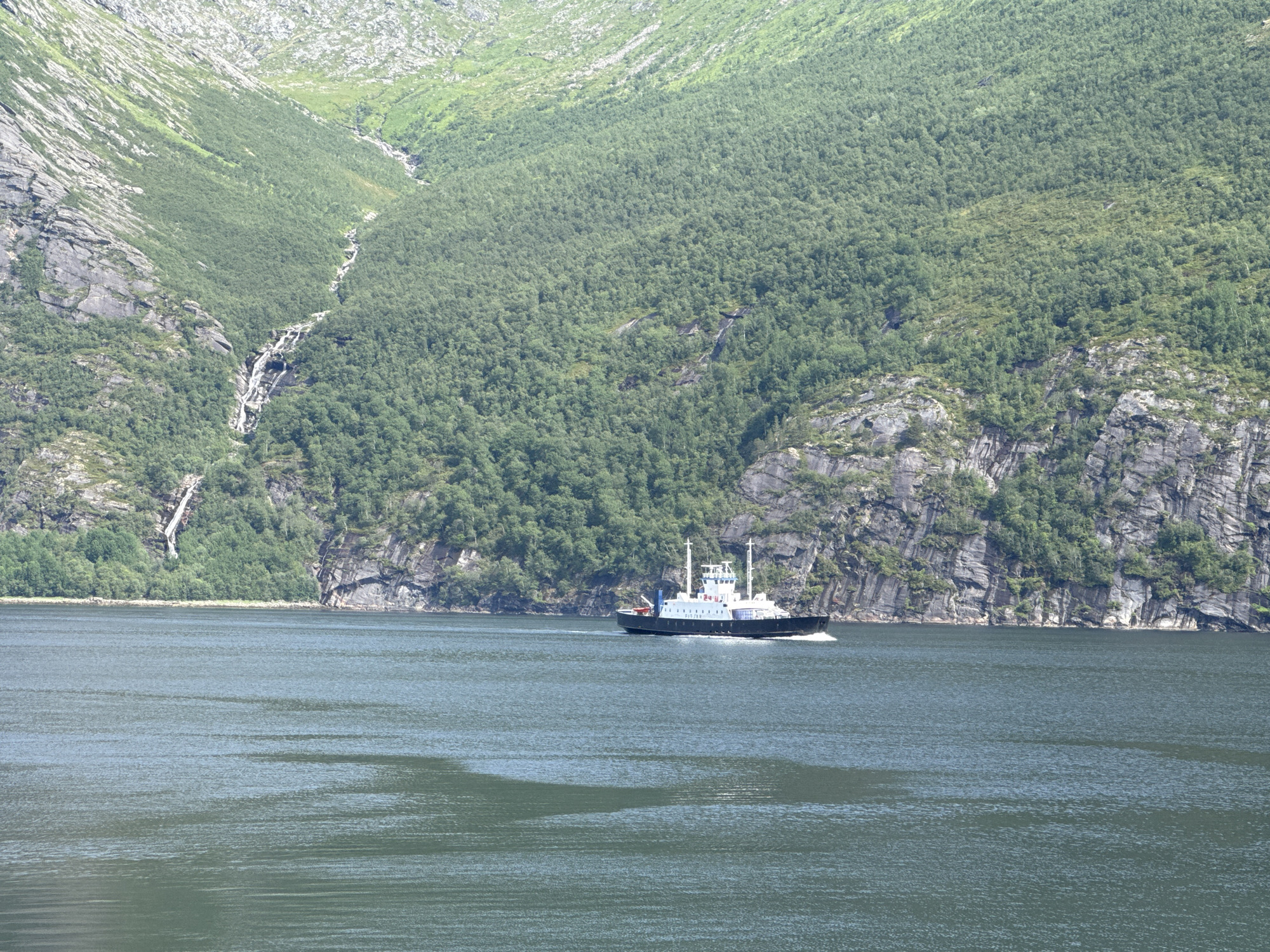 A ferry boat travels on a calm, dark green body of water. The ferry is white with a dark hull and has several decks and a white superstructure. It is moving from left to right. In the background, a steep, lush green mountainside rises dramatically from the water. A narrow waterfall cascades down the rocky, sparsely vegetated upper portion of the mountain. The lower slopes of the mountain are densely covered in green trees. The sky is not visible in the frame. The scene depicts a natural landscape with a man-made vessel. The location is identified as Inndyr, Norway.