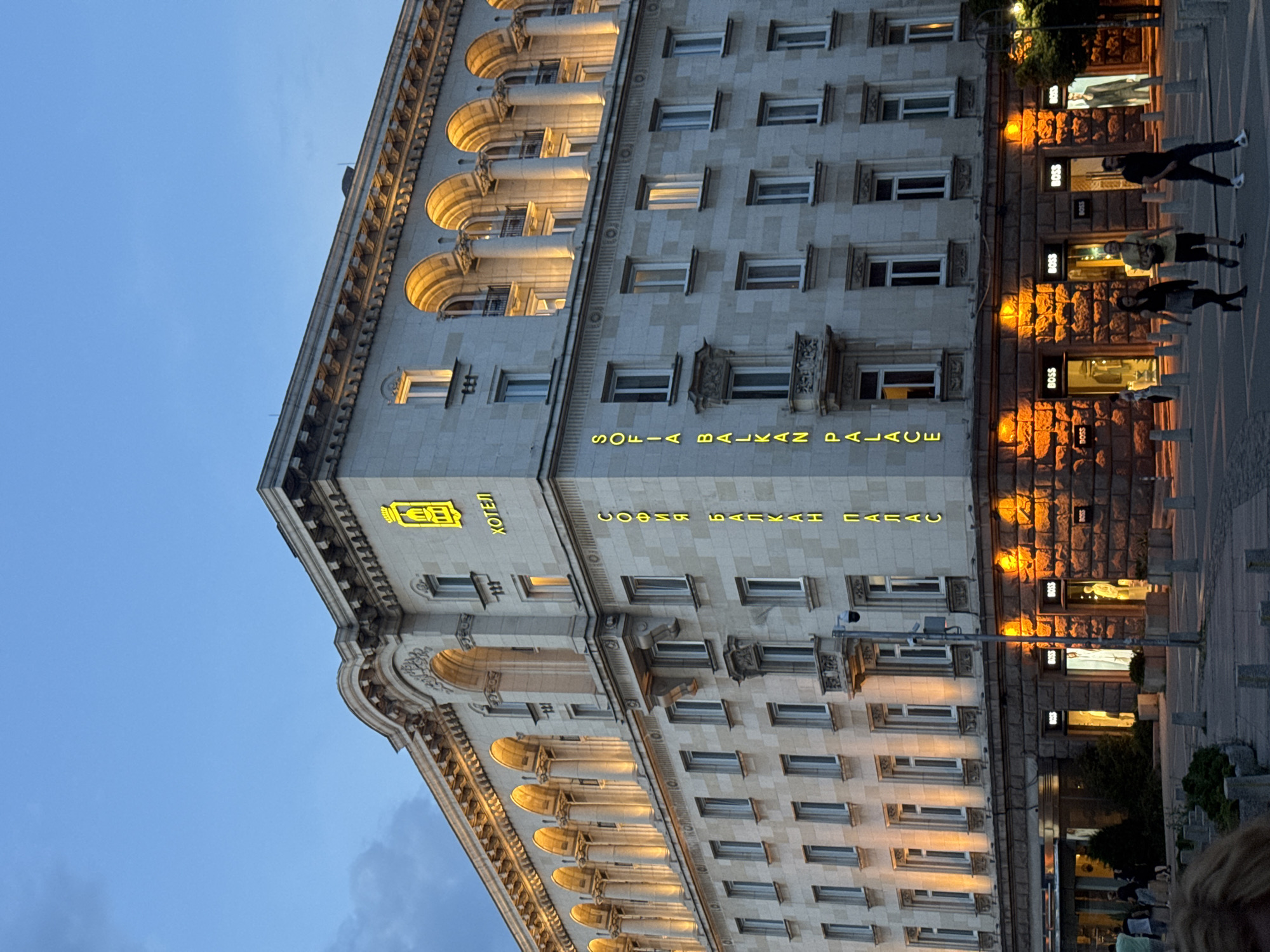 The image shows a nighttime exterior view of the Sofia Balkan Palace Hotel in Sofia, Bulgaria. The hotel is a large, ornate building with a light-colored stone facade. It features multiple floors with arched windows, and decorative architectural elements. Prominent yellow neon signs on the building read "ХОТЕЛ" (Hotel) and "СОФИЯ БАЛКАН ПАЛАС" (Sofia Balkan Palace). The lower level of the building houses several retail stores, identifiable by "BOSS" logos above their entrances. The street in front of the hotel is paved, and several pedestrians are visible walking across it. Streetlights and the illuminated building cast warm light onto the scene. The sky is a deep blue, indicating it is twilight or nighttime.