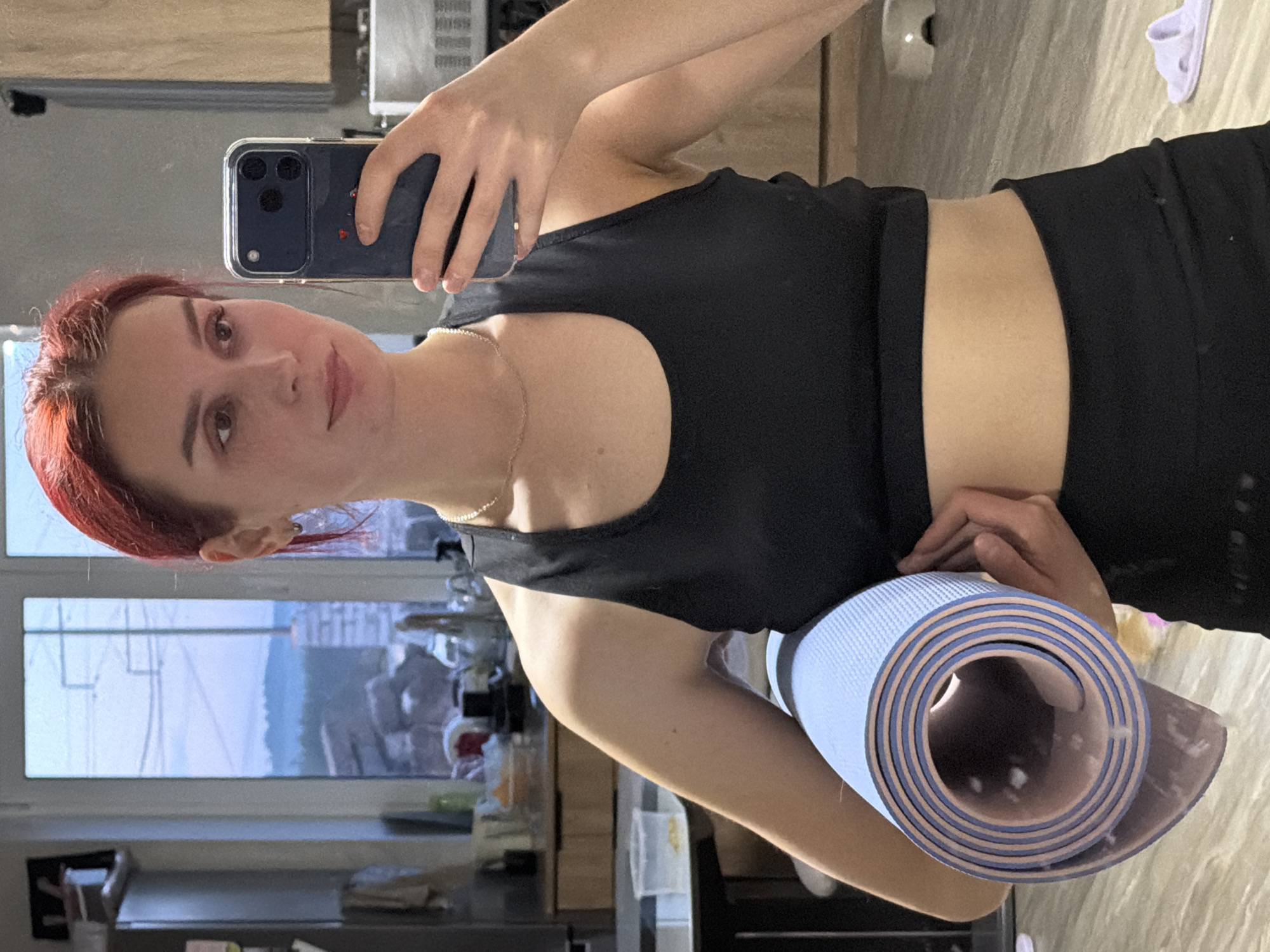 A young woman with reddish-brown hair, styled away from her face, stands indoors, captured in a mirror selfie. She wears a black sports bra and black leggings. Her expression is neutral, and she gazes directly towards the camera's reflection. Around her neck, she wears a delicate gold-colored chain, and a small earring is visible in her left ear.
She holds a dark-colored smartphone, likely an iPhone, in her right hand, positioned to take the picture. The phone is encased in a clear cover, which features a small red heart sticker on its back. In her left arm, she cradles a rolled-up yoga or exercise mat, which is light blue on its outer surface and light pink/purple on its inner side.
The background, visible through the mirror's reflection, suggests a home environment. A large window behind her reveals a bright daytime view of an urban landscape, including distant buildings, outlines of trees, and a light sky, consistent with the location context of Chaikovsky, Russia. Inside, reflections show what appears to be a kitchen or living area, with elements like a microwave oven, various items on shelves or counters, and light-colored slippers visible on the floor in the upper right portion of the image. The scene implies she is either preparing for or has just completed a fitness activity, given her attire and the yoga mat.