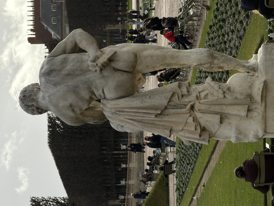 The image captures a scene in the Jardin du Luxembourg, Paris, France. The primary subject in the foreground is a large, weathered white marble statue of a muscular male figure viewed from the back. The figure is largely nude, with drapery partially covering his left thigh and lower torso. He holds a short, rectangular object, resembling a blade or tablet, behind his back with his left hand. The statue rests on a rough-hewn stone base, part of which is visible.
In the midground, beyond the statue, a public garden extends. Green lawns are visible, some sections containing rows of small, dark plants or dormant groundcover. A narrow dirt pathway traverses the lawn. Numerous lightweight green metal chairs are distributed across the grass and along pathways; many are occupied by individuals or small groups. People, dressed in jackets and coats, are observed sitting, relaxing, or walking. In the immediate lower-left foreground, a person in a dark jacket is seated on a green chair, facing away from the camera. A small white bird, possibly a seagull, stands on the grass to the left.
The background features a long, dark, densely pruned hedge or row of trees, forming a significant horizontal line. Above and behind this hedge, architectural elements of the Luxembourg Palace are visible, including a reddish-brown brick structure with a crenellated upper edge and a grey slate roof with dormer windows. The sky is partly cloudy with diffuse illumination.