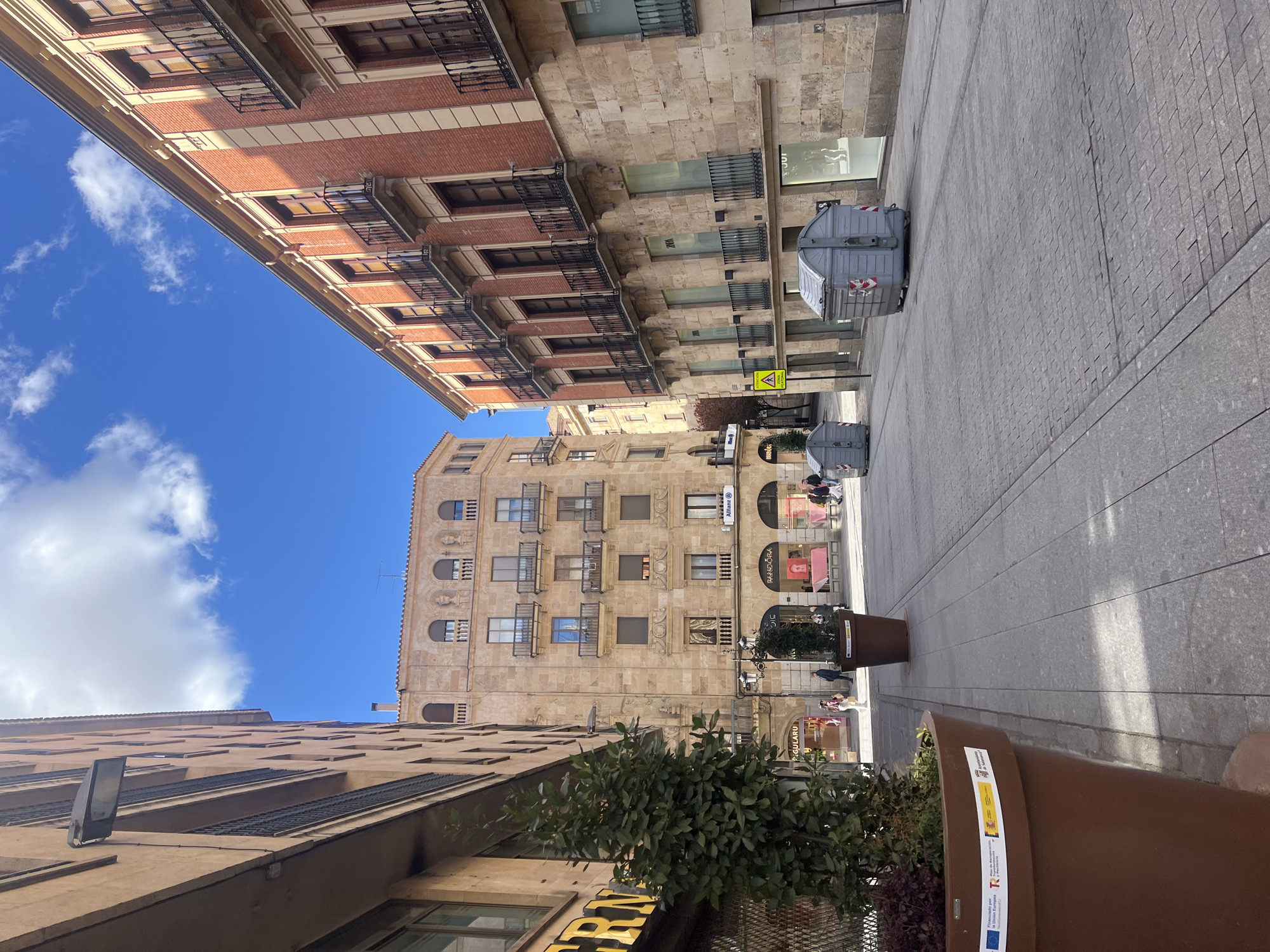 The image captures a street scene in Salamanca, Spain, looking up between buildings towards a blue sky with scattered clouds. The street, paved in gray stone, is bordered by a mix of architecture. Tall buildings line the sides of the street, featuring brick facades with balconies and stone structures with multiple windows.
A trash container is visible on the right side of the street. Sidewalk planters with foliage are along the edge of the pavement. Pedestrians can be seen further down the street, suggesting a bustling urban environment. The image is taken from a low angle, emphasizing the height of the buildings and the openness of the sky.