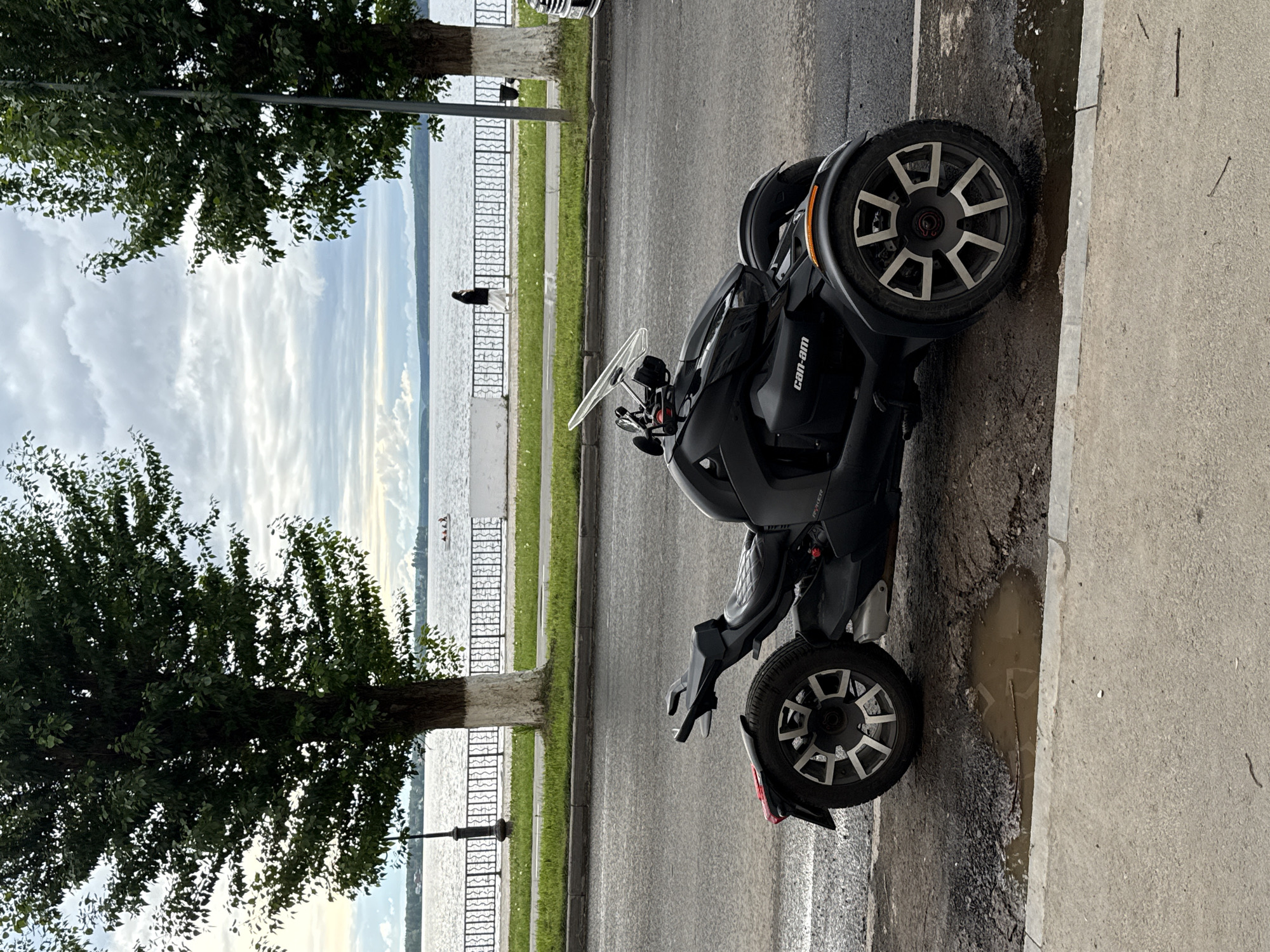 A black Can-Am Ryker three-wheeled motorcycle is parked on the right side of a paved road in Votkinsk, Russia. The road runs alongside a body of water, likely the Votkinsk Reservoir, which is visible in the background. A decorative metal fence separates the road from a grassy area along the waterfront. Trees line the side of the road opposite the motorcycle. On the far side of the decorative fence, several people are standing or walking along the waterfront. The sky is overcast with clouds. The curb and adjacent sidewalk are visible on the right side of the motorcycle. There is a puddle alongside the curb near the rear wheel of the motorcycle.