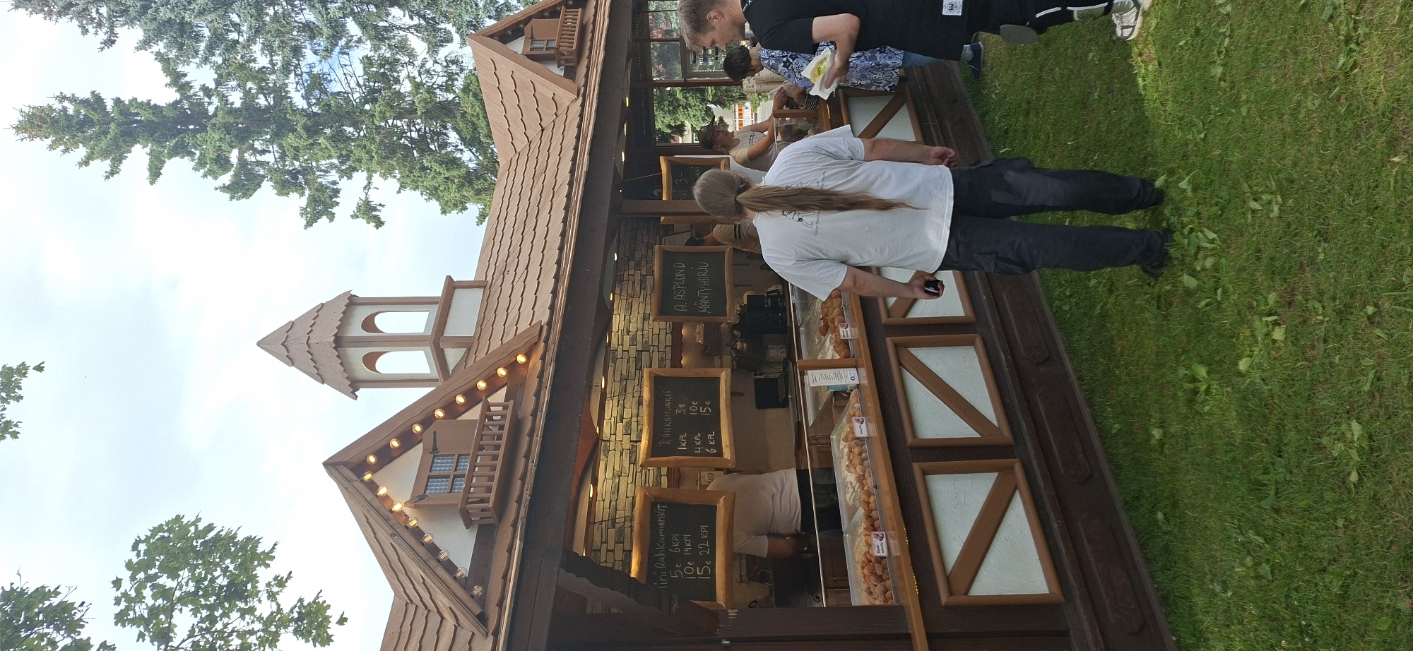In Turku, Finland, an outdoor food stall with a rustic, wooden facade displays baked goods. Three chalkboards list prices for items like "mini Rahkamunkit" and "Rahkamunkit" ranging from 3€ to 22€. Trays of pastries are visible behind a glass counter. Several individuals are present: a man with long hair in a ponytail is standing in the foreground, facing away from the camera. To his right, a woman holding a small white bag and a man wearing a black t-shirt are interacting. Another person is visible behind the counter. The stall is situated on a grassy area, with trees and a cloudy sky in the background. The architectural style of the stall suggests it might be a temporary structure, possibly for a festival or market.