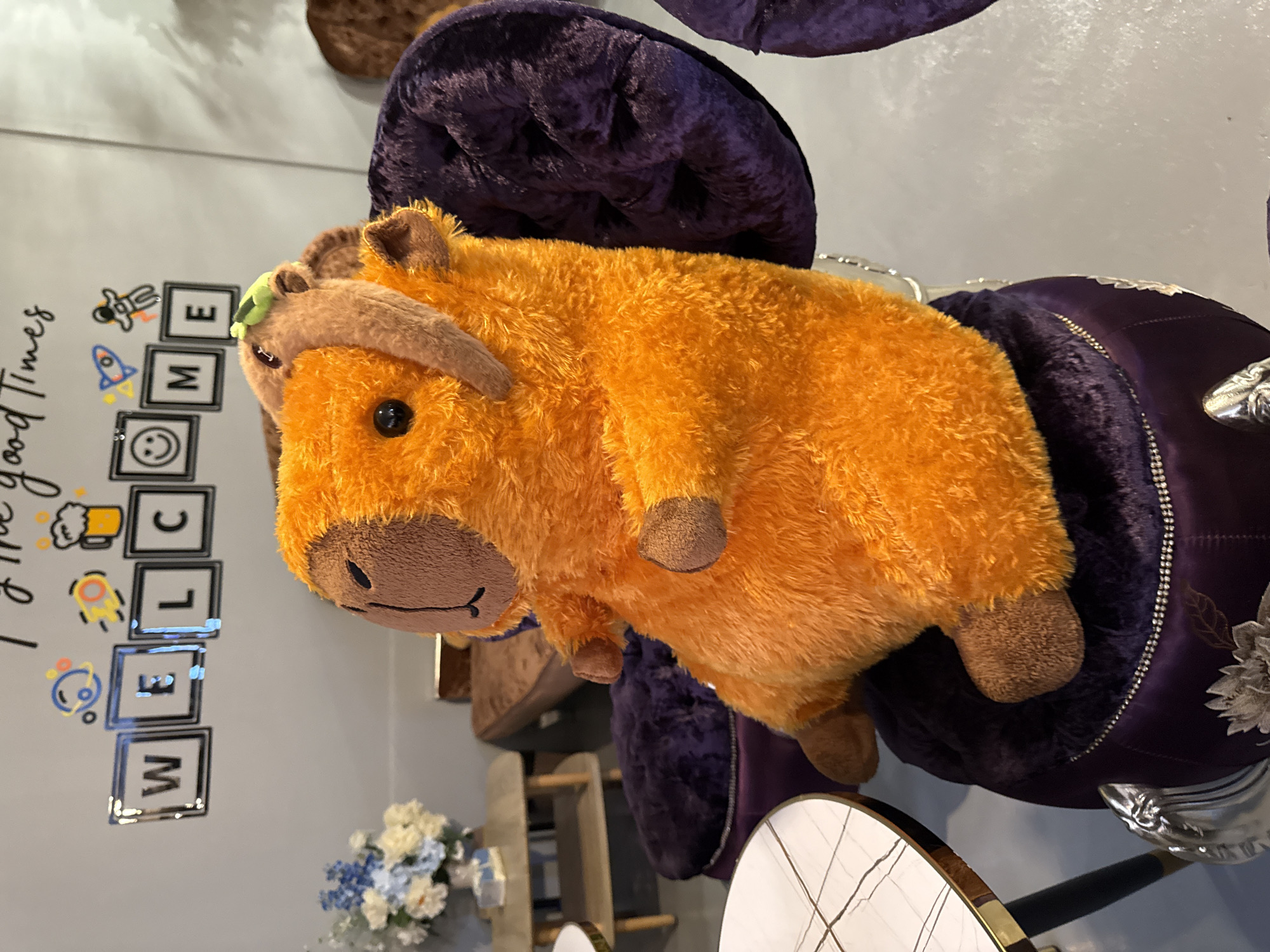 This image features a plush toy of an orange, cow-like creature with brown accents, possibly a Highland cow. The toy is resting on a plush, dark purple, circular seat, which appears to be part of a chair or ottoman, embellished with silver trim and floral embroidery. The toy is positioned in a way that its head is towards the bottom left of the frame, and its body extends upwards and to the right.
In the background, to the left of the toy, is a light gray wall with some decorative elements. These include the words "Good Times" written in cursive, and a series of square frames containing stylized icons. One frame has a figure in a spacesuit, another a rocket ship, a smiley face, and a few others that are difficult to discern. Below these, there are also letters spelling out "WELCOME" vertically, each in its own square frame, with some of the letters slightly obscured. A small bouquet of white and blue flowers is visible in the lower right corner of the image. The overall setting appears to be indoors, possibly in a home or a commercial establishment like a cafe or lounge, given the decorative elements and furniture. There are no humans visible in the image. The lighting suggests it's daytime, but no specific time can be determined. The scene evokes a sense of comfort and perhaps whimsical decor.