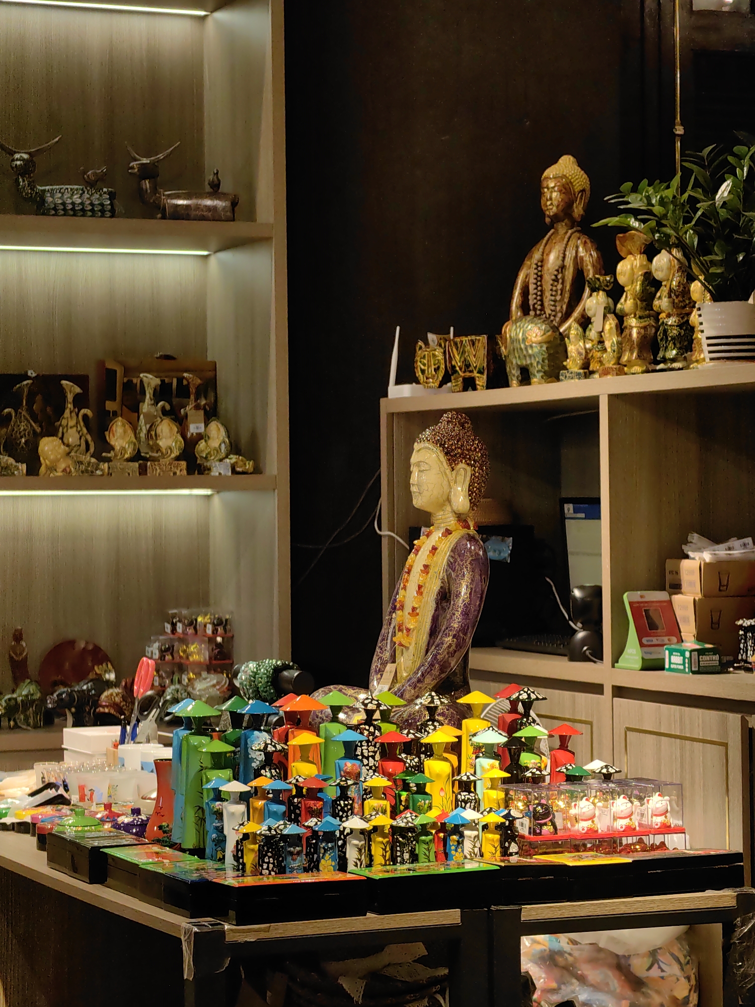 This media file captures the interior of what appears to be a gift or souvenir shop in Bai Chay Ward, Vietnam, showcasing a variety of decorative and cultural items. The setting is well-lit by artificial lights, including LED strips embedded in the wooden shelves on the left.
In the center foreground, a lighter-colored Buddha bust with a beaded topknot and a purple garment adorned with a yellow and orange beaded necklace is prominently displayed. To its right, on an upper shelf, sits another, larger golden-brown Buddha statue, flanked by a small green elephant figurine and other golden sculptures, alongside a potted green plant.
The left side of the image features two tiers of light wood shelves, brightly lit. The top shelf holds two dark, ornate animal figures resembling deer, accompanied by small bird-like sculptures. The shelf below displays an assortment of intricately shaped vases and other abstract decorative pieces.
The main display table in the foreground is vibrant with color. It features numerous small, stylized figurines arranged in rows, characterized by their bright hues (blue, green, yellow, orange, black) and distinctive conical hats, some with patterned bodies. Next to these, several clear plastic boxes hold white and red Maneki-neko (beckoning cat) figurines. Other items on the table include red scissors and various trinkets.
On the shelves to the right, behind the central Buddha bust, are a computer screen, various packaged goods, and a green box with the visible text "CONTRO" and "ROBBO". Another box partially visible above it reads "VIC 16". The overall impression is one of an inviting retail space filled with artisan crafts and souvenirs, reflecting local culture, arranged for display and sale.