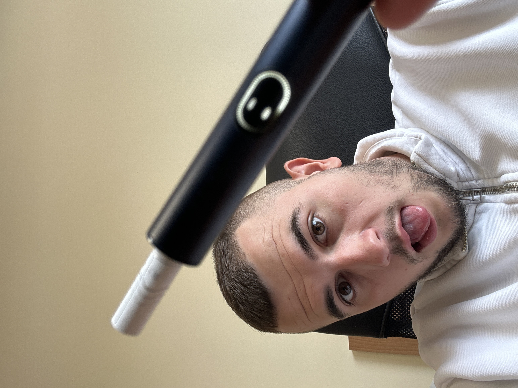 A selfie is taken indoors, with the photographer holding a black, electric toothbrush with a white tip towards the camera. The person is a light-skinned man with a beard. He is wearing a white hoodie and has his tongue sticking out. The background is a beige-colored wall and a black office chair. It is not possible to determine the exact location within Plovdiv, Bulgaria, based on this image alone.