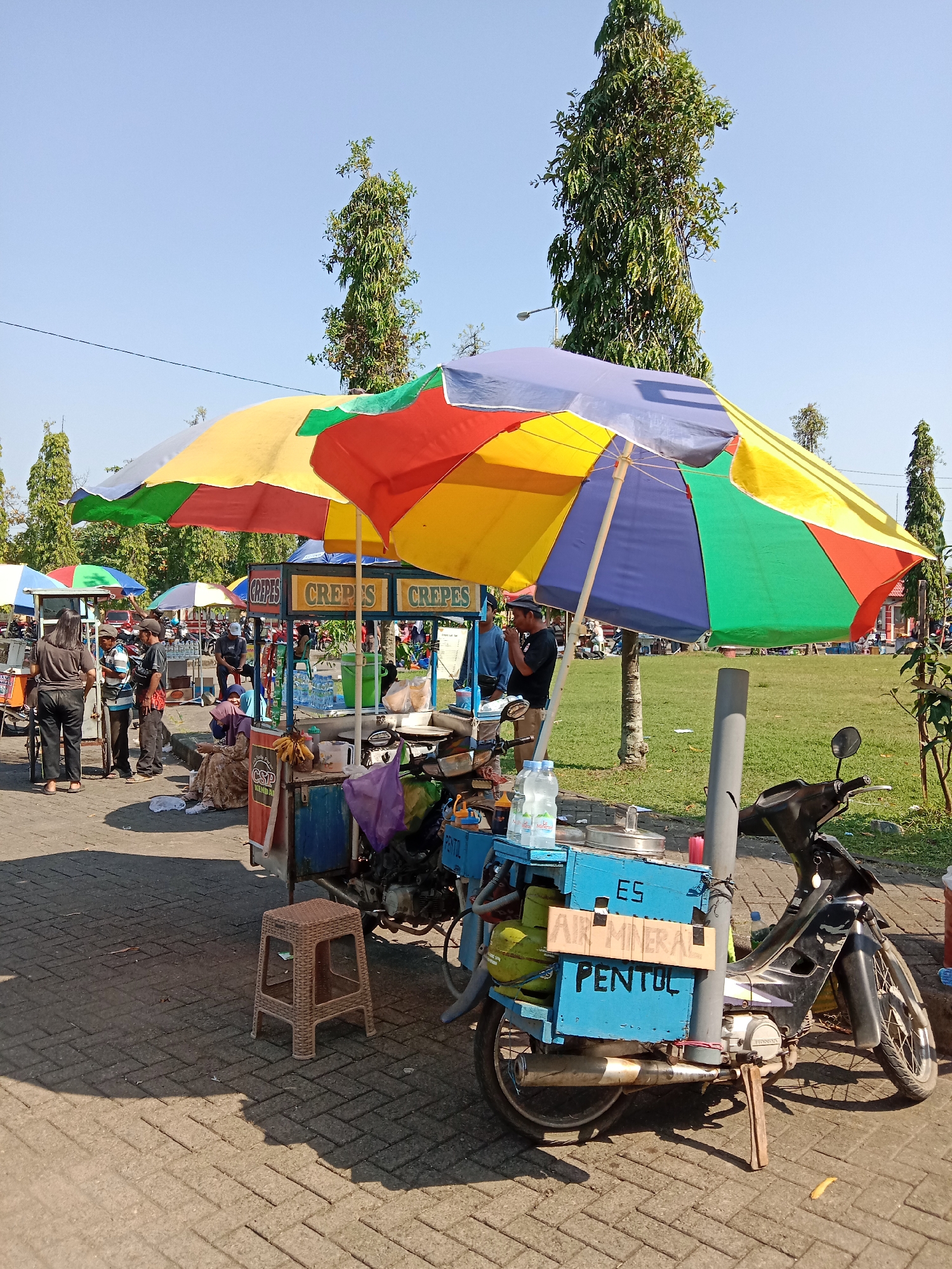 The image depicts a street scene in Kendal, Indonesia, likely during the day given the bright sunlight and clear blue sky. In the foreground, a mobile food cart is set up, featuring a large, colorful umbrella providing shade over a display of goods. The cart is attached to a motorcycle, suggesting it is a portable vendor. Signs on the cart indicate items for sale, including "Crepes," "Es Air Mineral," and "Pentul," which are Indonesian snacks and drinks. Several people are visible, some appearing to be customers and others vendors. One woman in a brown headscarf is seated near the cart, while others stand and interact. In the background, more food carts and umbrellas are visible, along with trees and a grassy area, suggesting this is a public space or market area. The ground is paved with interlocking bricks.