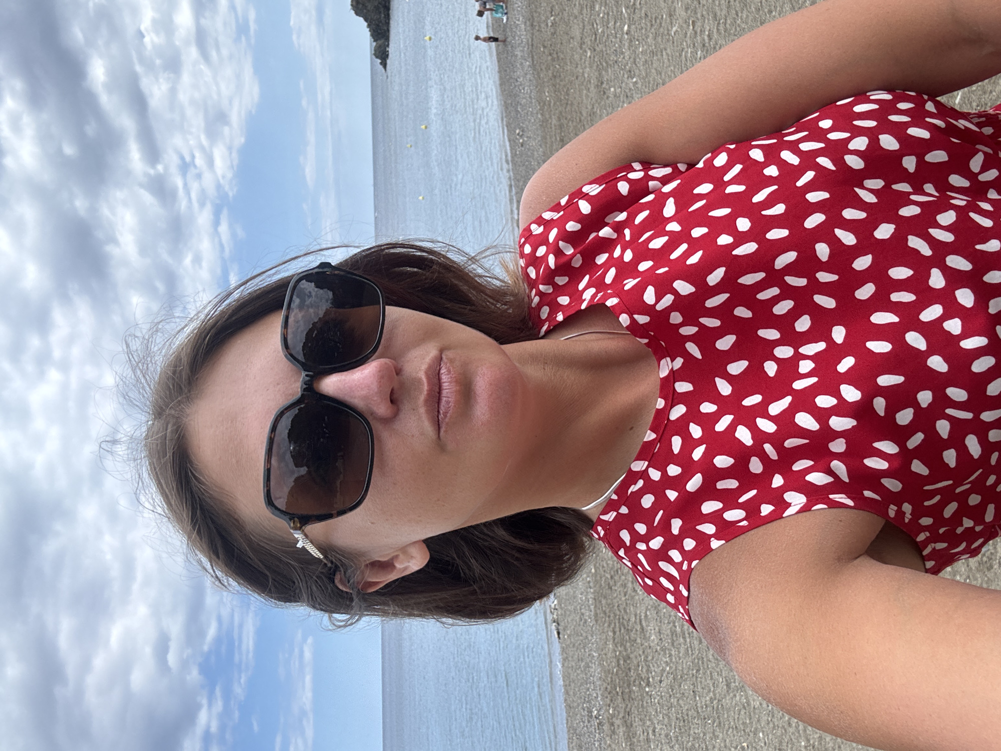 A woman is posing for a selfie on a beach in Port-Vendres, France. She is wearing large, dark sunglasses and a red dress with white polka dots. The beach is covered in pebbles and sand, and the ocean is visible in the background. There are a few people in the distance near the water. The sky is cloudy.