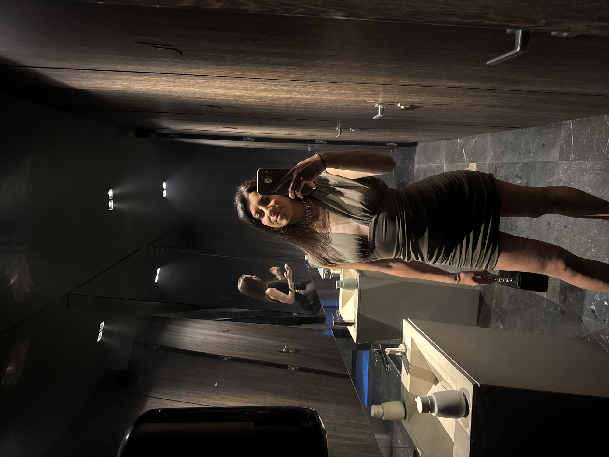 A woman is taking a selfie in a bathroom. She is standing in front of a mirror and holding a smartphone in her right hand. She is wearing a short dress and necklace. The bathroom has a modern design with a dark color scheme, wooden partitions, and stone floors. There are several sinks with modern faucets. It is not possible to confirm the exact location in Blagoevgrad, Bulgaria from the image.