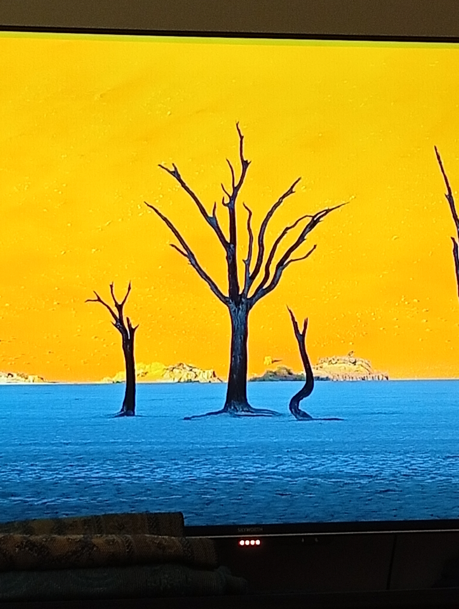 The image depicts a stark landscape with a bright yellow sky and a vast expanse of blue water. In the foreground, three silhouetted, leafless trees stand in the water, their branches reaching towards the sky. The largest tree is in the center, with two smaller trees to its left and right. In the background, a distant, rocky landmass is visible. The overall impression is one of desolation and beauty. There are no people visible in the image, and no specific details can be ascertained about the location in Төмөнкү Ала-Арча, Kyrgyzstan, as the image appears to be a generalized landscape rather than a specific geographical landmark.
