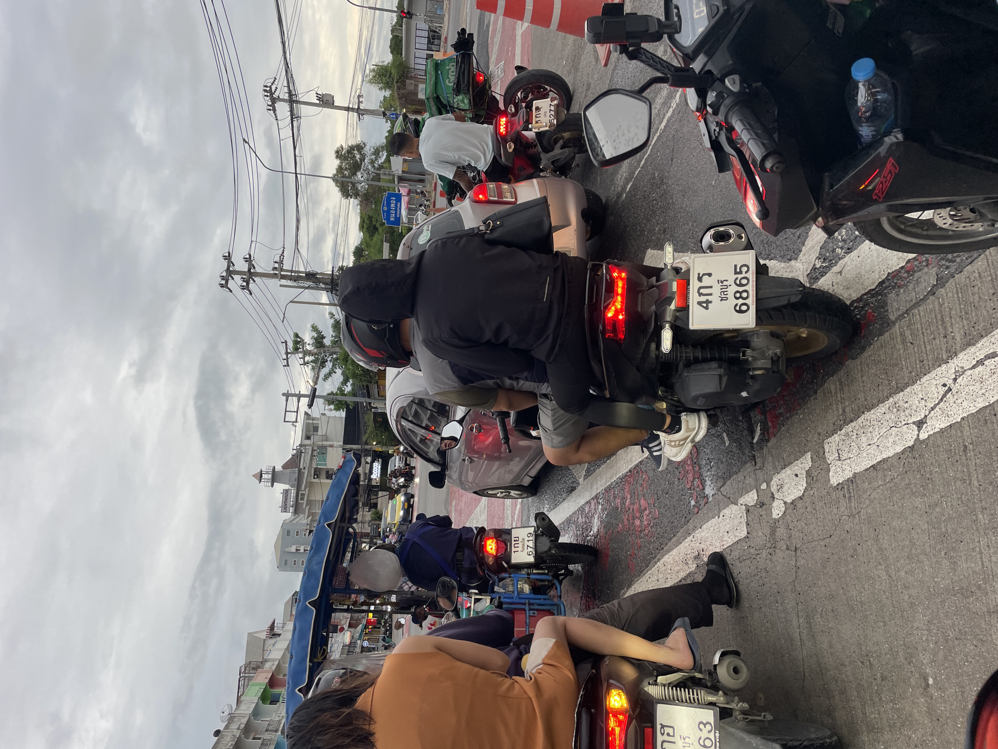 The image captures a traffic scene in Pattaya City, Thailand, likely during a period of congestion. Several motorcycles and a car are visible, all appearing to be stationary or moving at a very slow pace. The sky is overcast, suggesting a cloudy or potentially rainy day.
In the foreground, a person wearing an orange top is seated on a motorcycle, facing away from the camera. To their left, another motorcycle is also stopped. The motorcycle immediately behind the person in the orange top has a license plate with Thai script and the number "6719". This motorcycle is occupied by two individuals; the rear passenger is wearing a dark hooded jacket.
To the right of the central motorcycle, a light-colored car is stopped, and to the right of the car, another motorcycle is visible with a rider wearing a white t-shirt and a green helmet. This motorcycle has a green delivery box attached to the back, with the "GRAB" logo clearly displayed, indicating a delivery service.
Further back in the scene, other vehicles, including cars and motorcycles, are visible in the traffic. Buildings line the street, with some bearing signage and awnings. Electrical wires and utility poles are prominent overhead, typical of urban infrastructure. The road surface appears wet in places, with painted white lines and what seem to be red markings. The overall impression is of a typical urban traffic environment in Pattaya City.