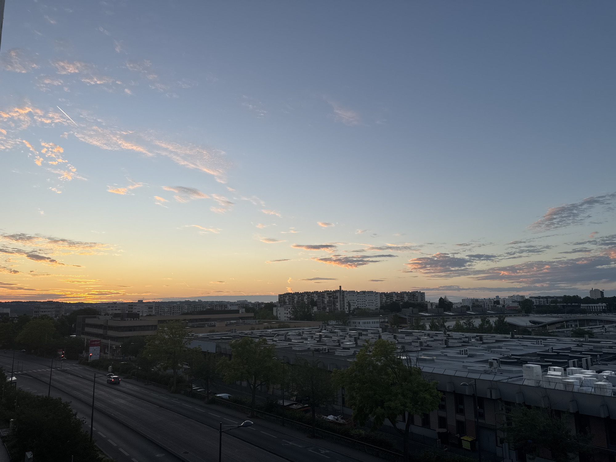 The image shows a city skyline at sunset. The sky is a mix of orange, pink, and blue, with scattered clouds. There is a contrail from an airplane visible in the sky. In the foreground, there is a multi-lane road with cars driving on it. The road is lined with trees and buildings. In the background, there are taller buildings and apartment complexes. The overall scene depicts an urban environment at dusk.