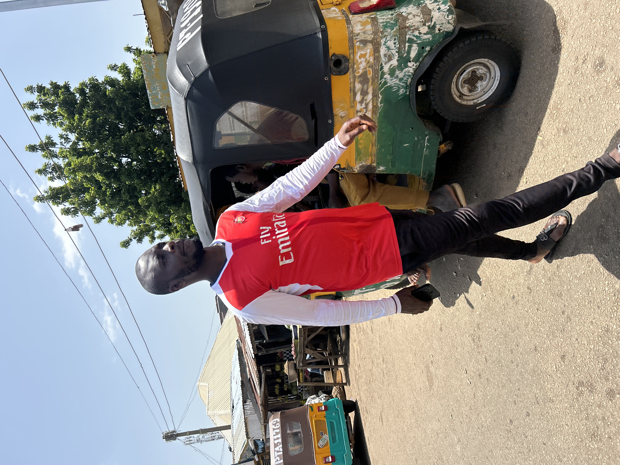 In Jos, Nigeria, a man wearing a red and white long-sleeved shirt, possibly a sports jersey, with "Fly Emirates" printed on it, walks alongside a multi-colored auto-rickshaw, or Keke Napep. The Keke, predominantly yellow and green with areas of rust and peeling paint, is parked on a paved street. Inside the Keke, at least one other person is visible. In the background, market stalls with tin roofs and various goods, likely produce, line the street. Power lines and a large green tree are also present in the scene, under a bright, clear sky. The man is holding a dark-colored mobile phone in his right hand.