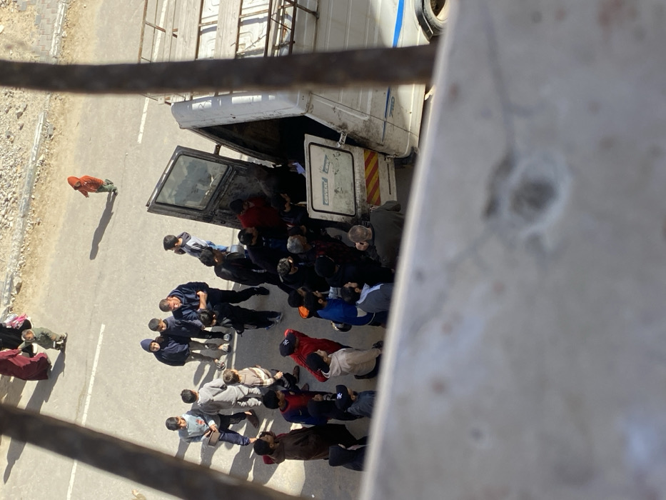 An overhead photograph depicts a street scene in Khan Yunis, Palestinian Territory, under bright daylight with strong shadows. The immediate foreground shows an out-of-focus concrete ledge or railing with a metal bar on the right.
On the paved street, a white van, marked "COVERT LORRY" and "710" with yellow and red striped hazard tape on its lower side panel, is stationary. Its passenger-side front door is open, and its rear or large side cargo doors are also open, facing a gathering crowd.
Approximately 20-30 individuals, predominantly men and boys, are clustered densely around the open cargo area of the van. Several individuals appear to be leaning into or interacting with the van's interior. Other children and adult males are standing nearby, observing or waiting. One small child in a red hooded jacket walks alone on the street further from the main group, casting a long shadow. In the upper left background, a woman in a maroon abaya and head covering is walking with another small child. The street is asphalt, bordered by a dirt and rubble shoulder on the right.