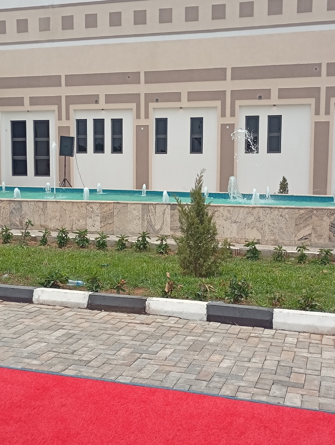 This media file captures the exterior of a modern building and its surrounding landscaped area, located in Unguwan Sanusi, Nigeria. The building's facade is characterized by a two-tone design, featuring a beige upper section with decorative square elements and a lower part with white and beige rectangular panels framing dark-colored, tall rectangular windows.
In the foreground, a prominent water fountain runs horizontally across the frame, filled with clear, light blue-green water. Numerous small jets spray water upwards, with some larger, more energetic splashes visible, especially on the right side. The fountain basin is constructed from light-colored stone or tile.
Beyond the fountain, a neatly manicured lawn with green grass and several small bushes and shrubs is visible. A single, taller, conical evergreen plant stands out near the center. This green space is bordered by a curb painted with alternating black and white sections, separating it from a paved area made of interlocking grey and brown pavers.
A vibrant red carpet is laid out over the paved area in the foreground, extending from the bottom left upwards, strongly suggesting preparation for a formal event or ceremony. A black speaker on a tripod stand is positioned to the left of the fountain, reinforcing the likelihood of an organized gathering.
The scene is brightly lit by natural daylight, indicating daytime. No people are present, and no text is visible within the image. The overall atmosphere suggests a well-maintained venue ready for an occasion.