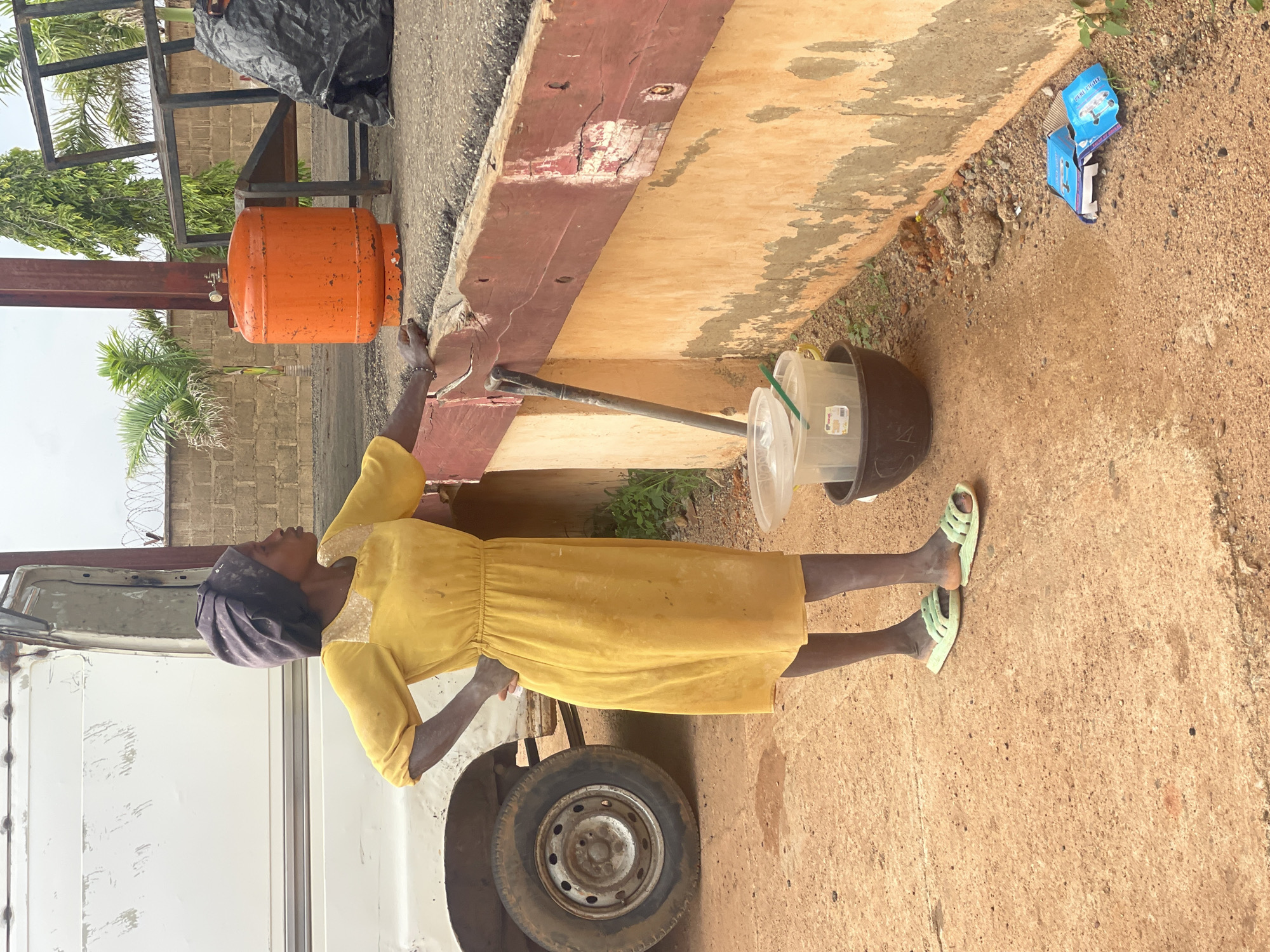 A medium shot captures a person in a yellow dress and a dark headscarf standing by a concrete wall in Kwang, Nigeria. The individual faces left, with their right hand resting on the wall. To the left, the front of a white utility vehicle is visible, including its tire. An orange gas cylinder stands to the right of the person, near a metal structure and a black plastic bag. A wooden plank, painted red, runs along the top of the concrete wall, showing signs of wear. Below the plank, there are two buckets, one clear plastic and one dark, with a green tool handle protruding from them. The ground is a mix of reddish-brown soil and gravel. In the lower right corner, two discarded blue boxes with white lettering are on the ground. The background includes a brick wall and greenery, suggesting an outdoor urban setting.