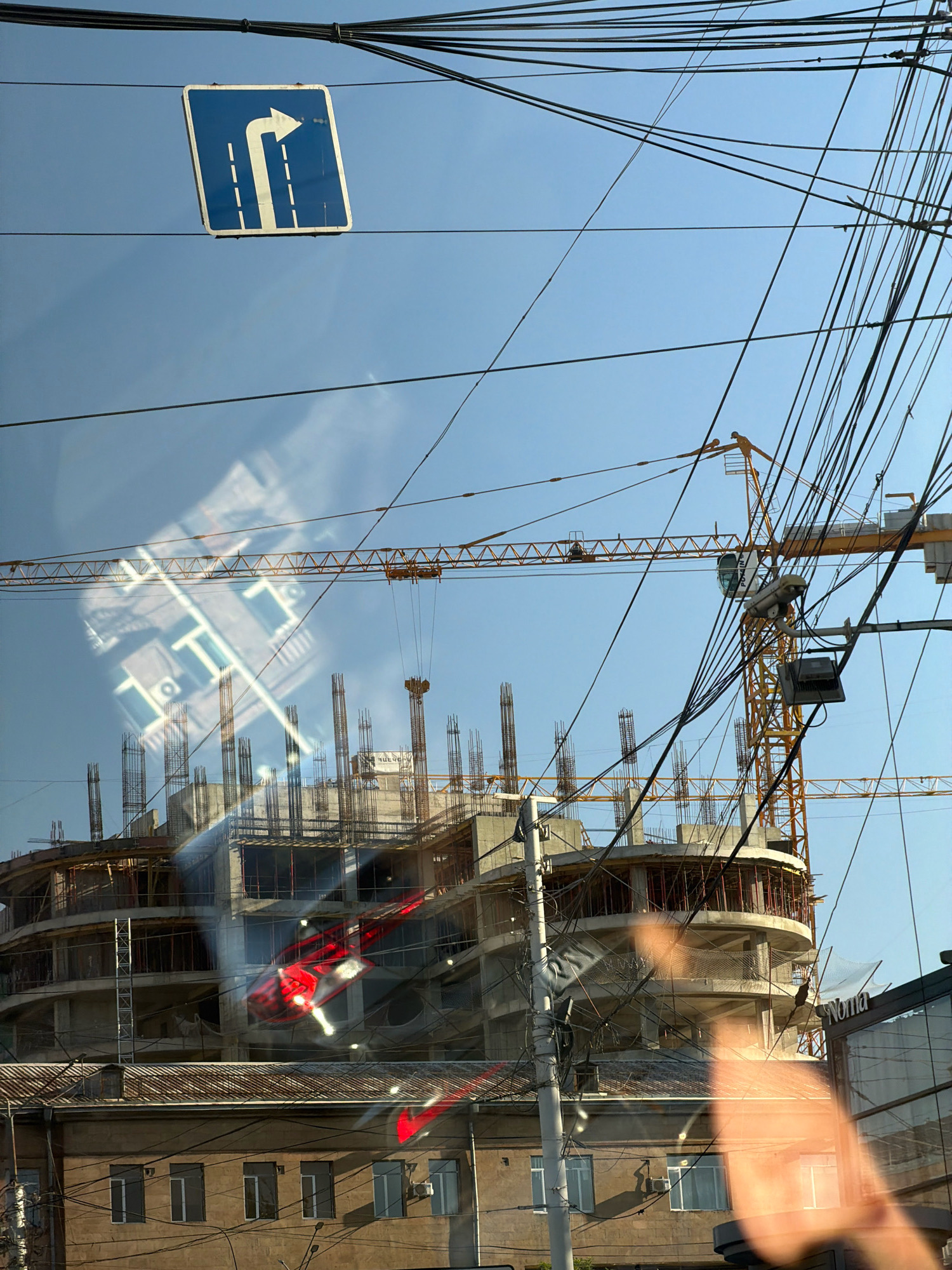 The image depicts a construction site in Yerevan, Armenia, with a partially built multi-story building dominating the background. Several yellow construction cranes are visible, with their long jibs extending across the frame. A complex network of overhead electrical wires crisscrosses the sky, obscuring parts of the scene. A blue and white road sign, indicating a mandatory right turn with two lanes, is mounted on a wire. Reflections on a glass surface, possibly from a window or vehicle, show abstract patterns and the shape of a building. Below the construction site, there is an older, multi-story building with numerous windows. The sky is clear and blue.