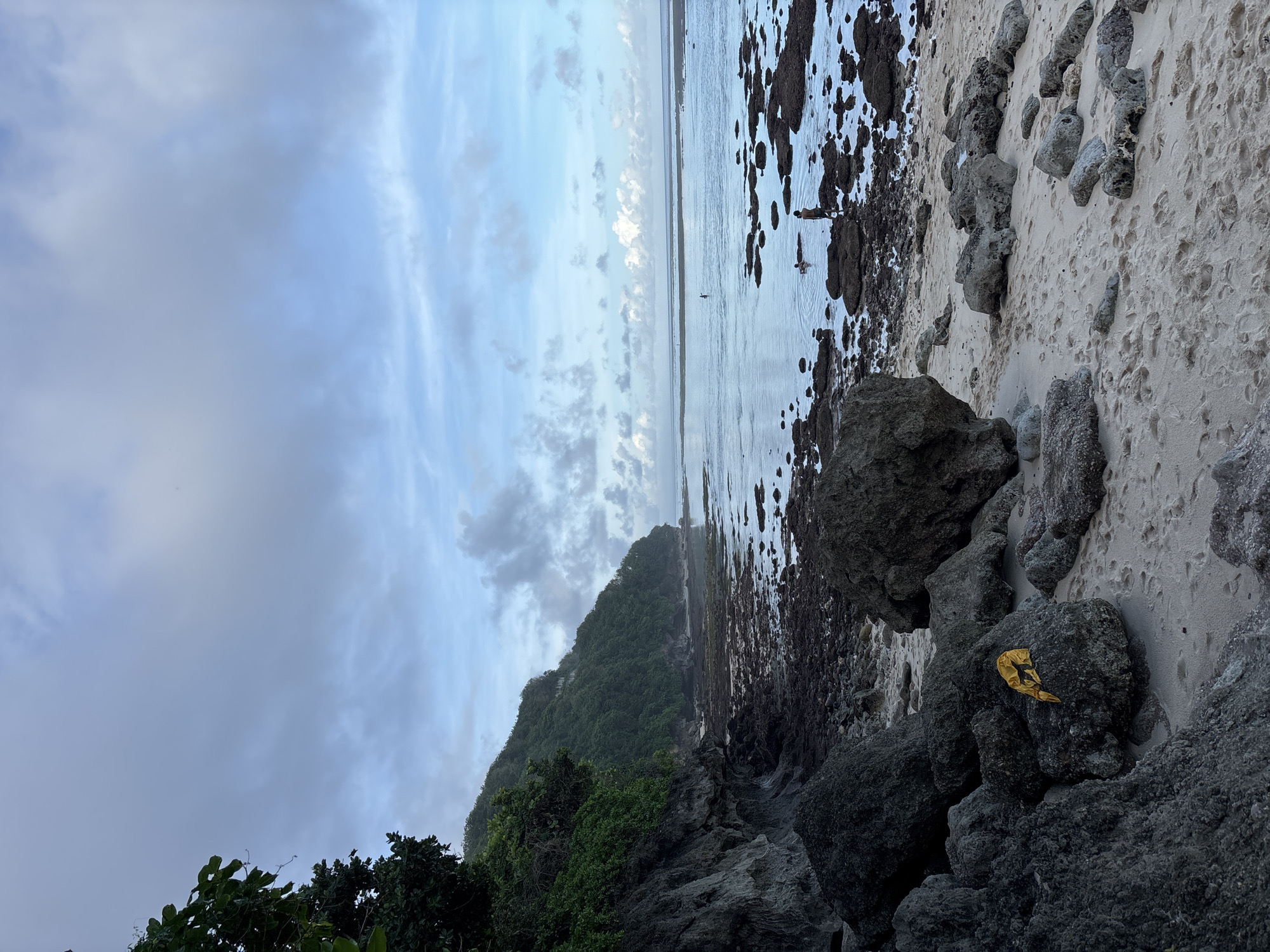 The image captures a coastal scene in Kuta Selatan, Indonesia. The sky is overcast with scattered clouds. A lush, green hillside slopes down towards a beach. The shoreline consists of a mix of white sand, rocky areas, and exposed seabed. Several large boulders are visible along the edge of the beach, with a discarded golden wrapper resting on one. In the water, two figures appear to be wading. The ocean stretches to the horizon, meeting the cloudy sky.
