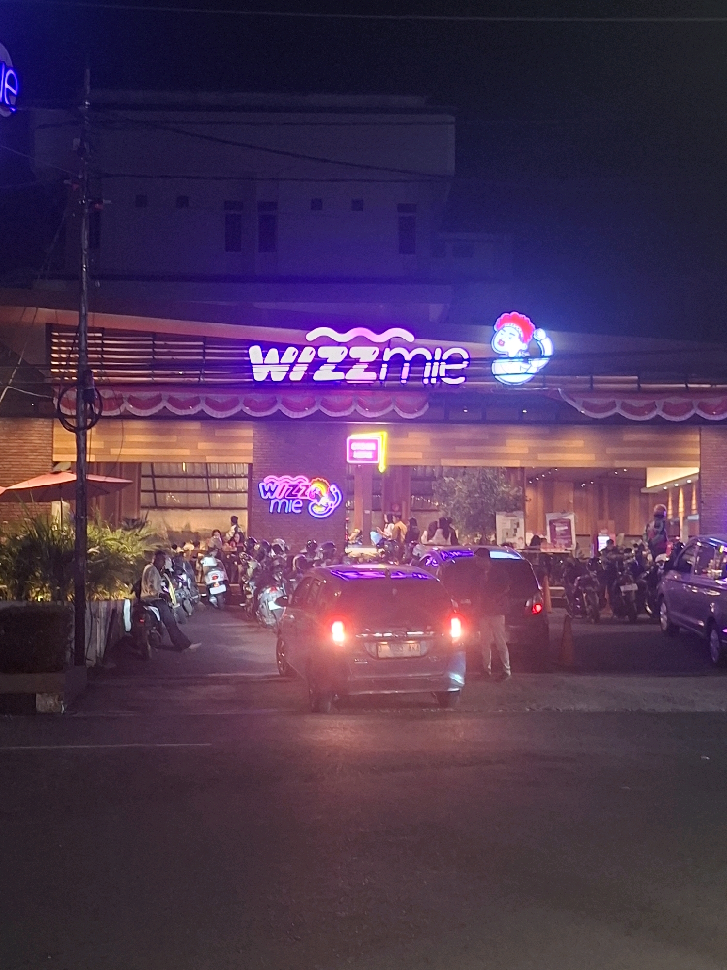 The image captures a nighttime scene outside a Wizzmie restaurant in Cianjur, Indonesia. The restaurant's facade is illuminated by purple neon signage that spells out "Wizzmie" in large, stylized letters, accompanied by a cartoon character. Red and white decorative bunting hangs above the entrance.
In front of the restaurant, a street is visible with several vehicles, including motorcycles parked in rows and cars on the road. A black car with its brake lights illuminated is in the foreground on the left, and another dark-colored car is to its right, also with brake lights on. People are seen standing and sitting near the parked motorcycles. The environment appears to be a commercial area, with the building lights and signage indicating a bustling establishment. The overall atmosphere is lively, typical of a restaurant scene at night.