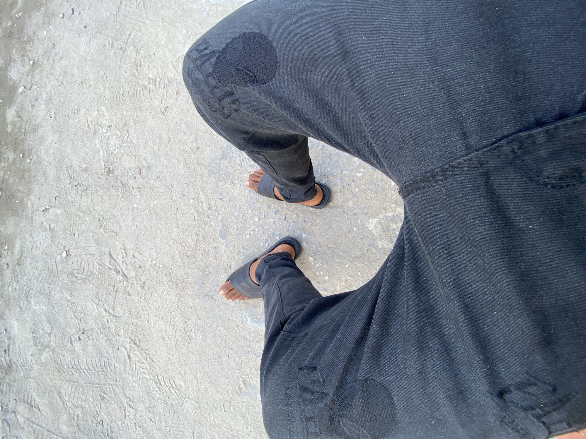 The image depicts a downward view of a person standing on a grey concrete surface. The individual is wearing dark-colored jeans and dark-colored sandals. The jeans have a circular patch or design on both legs, with text visible within the patch. The location is in Ado, Nigeria. The environment is an outdoor setting with natural light and a concrete surface exhibiting dirt and tire marks.