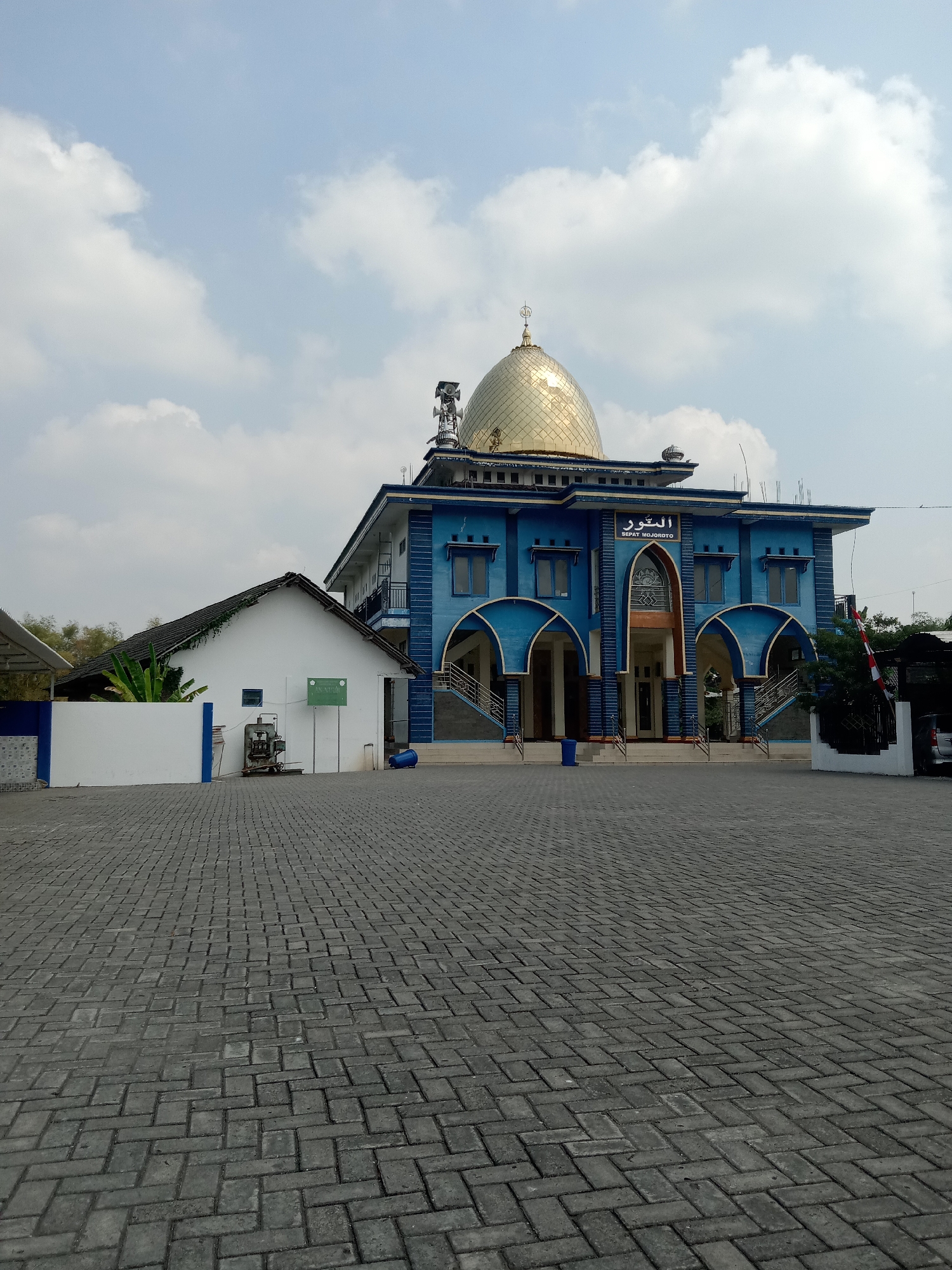 The image shows a blue mosque with a golden dome in what appears to be a semi-urban or suburban area. The mosque has an elaborate facade with arched entrances and decorative elements. A smaller white building with a dark roof is adjacent to the mosque. The ground in front of the buildings is paved with dark grey interlocking bricks. The sky is partly cloudy with patches of blue. A sign with Arabic script and the words "SIPAT MOJOROTO" is visible on the mosque. There are some utility poles and antennas on top of the mosque, and a few scattered objects on the ground including a blue barrel and what looks like a piece of machinery in front of the white building. There are no people visible in the image. The presence of the name "Mojoroto" on the sign suggests a potential connection to the Mojoroto area of Kediri, East Java, Indonesia. However, the prompt specifies the location as Sidobandung, Indonesia. Given that Mojoroto is a known place name in Indonesia, and without further context, it is difficult to definitively confirm the exact location as Sidobandung based solely on the visual information provided.