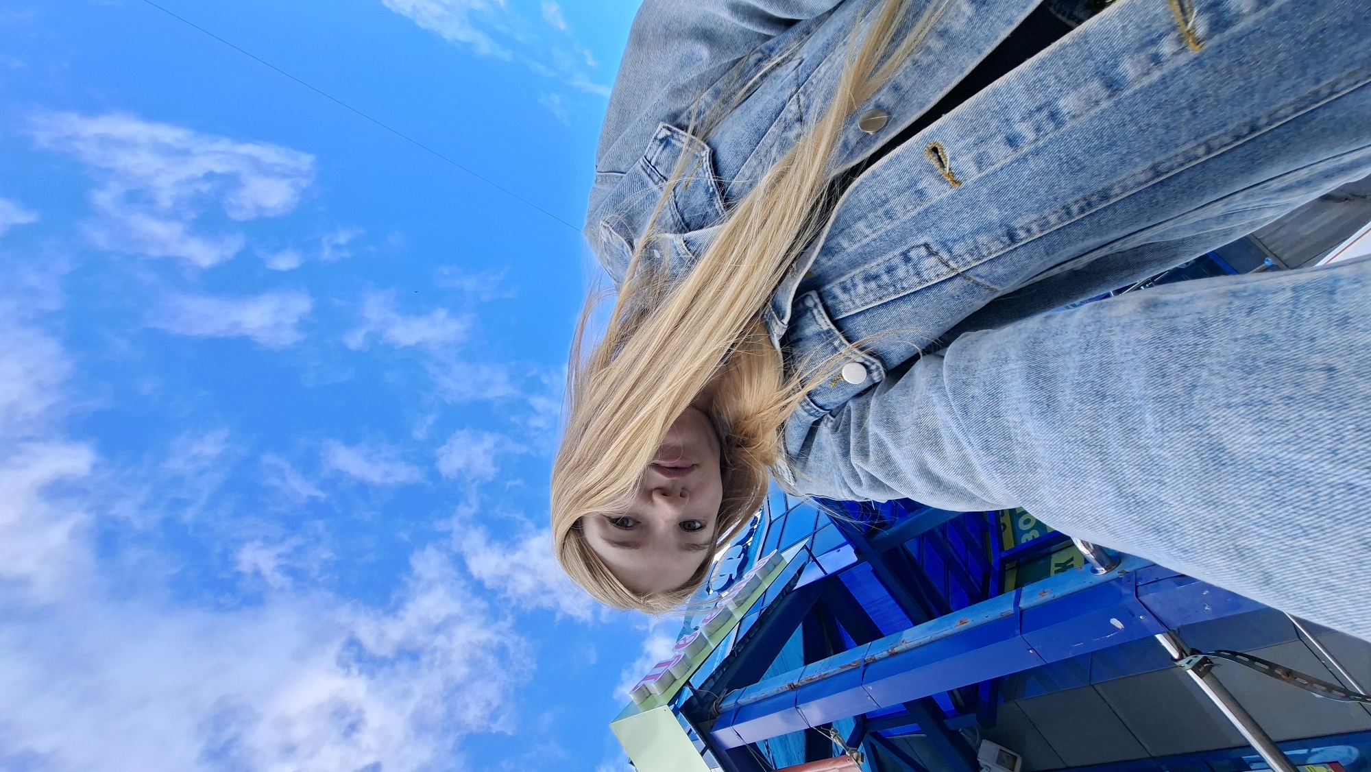 The image is a low-angle, slightly tilted selfie taken outdoors against a bright blue sky with scattered white clouds. In the foreground, a young woman with long blonde hair is wearing a light blue denim jacket. Her face is turned towards the camera, with her head tilted and her hair falling across her face. To the left of the woman, a blue and green structure with some signage is visible, appearing to be part of an amusement park ride or attraction. The composition suggests the photo was taken at an amusement park. The bright sky and the presence of an amusement park suggest an outdoor daytime setting. The specific location of Chaikovsky, Russia, cannot be determined from the visual information provided in the image.