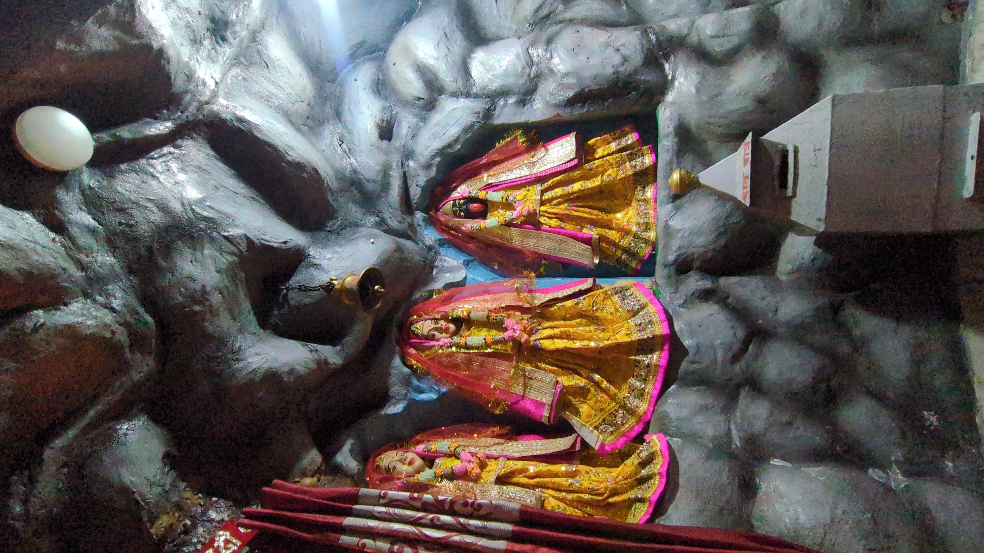 The image shows an interior view of a small Hindu shrine in Bhagsu, India. Three statues of deities, adorned in yellow and pink robes, are placed within recessed niches in a rough, cave-like wall. A small bell hangs from a chain to the left of the figures. A white dome-shaped object is attached to the wall further left. To the right of the statues is a gray, box-like structure with a gold-colored ornament. A patterned red cloth lies in front of the bottom figure. The walls are textured and painted gray, resembling a cave.