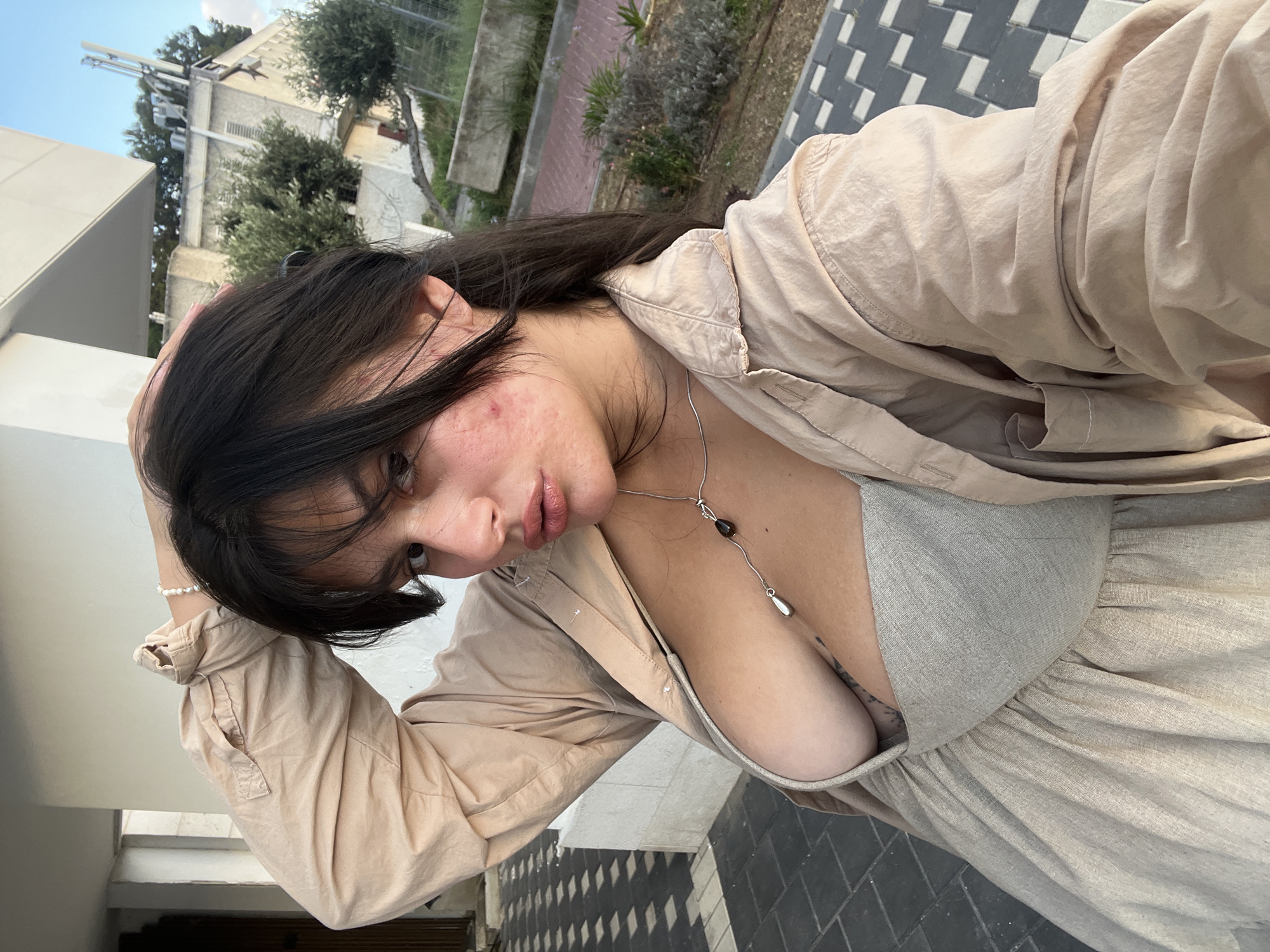 The media file shows a young woman with long, dark hair, taking a selfie outdoors. She is wearing a light-colored button-down shirt open over a grey top, and a flowing skirt of the same material. A delicate silver necklace with a dark pendant rests on her chest. Her hand is raised to her head, and she is looking down and to the side, with her mouth slightly open. Her skin shows some redness and blemishes, particularly on her cheek.
The background suggests an urban or suburban setting. In the upper left, a building with what appear to be communications equipment on its roof is visible, along with some trees. The ground is paved with a patterned tile. The lighting suggests it is daytime, possibly late afternoon or early evening, given the angle of the light and the shadows. The woman's expression is difficult to discern clearly due to the angle of the shot and her gaze, but it does not appear overtly happy or sad; perhaps thoughtful or relaxed. The overall scene is casual and personal. There is no visible text in the image. The location context is Kiryat Bialik, Israel.