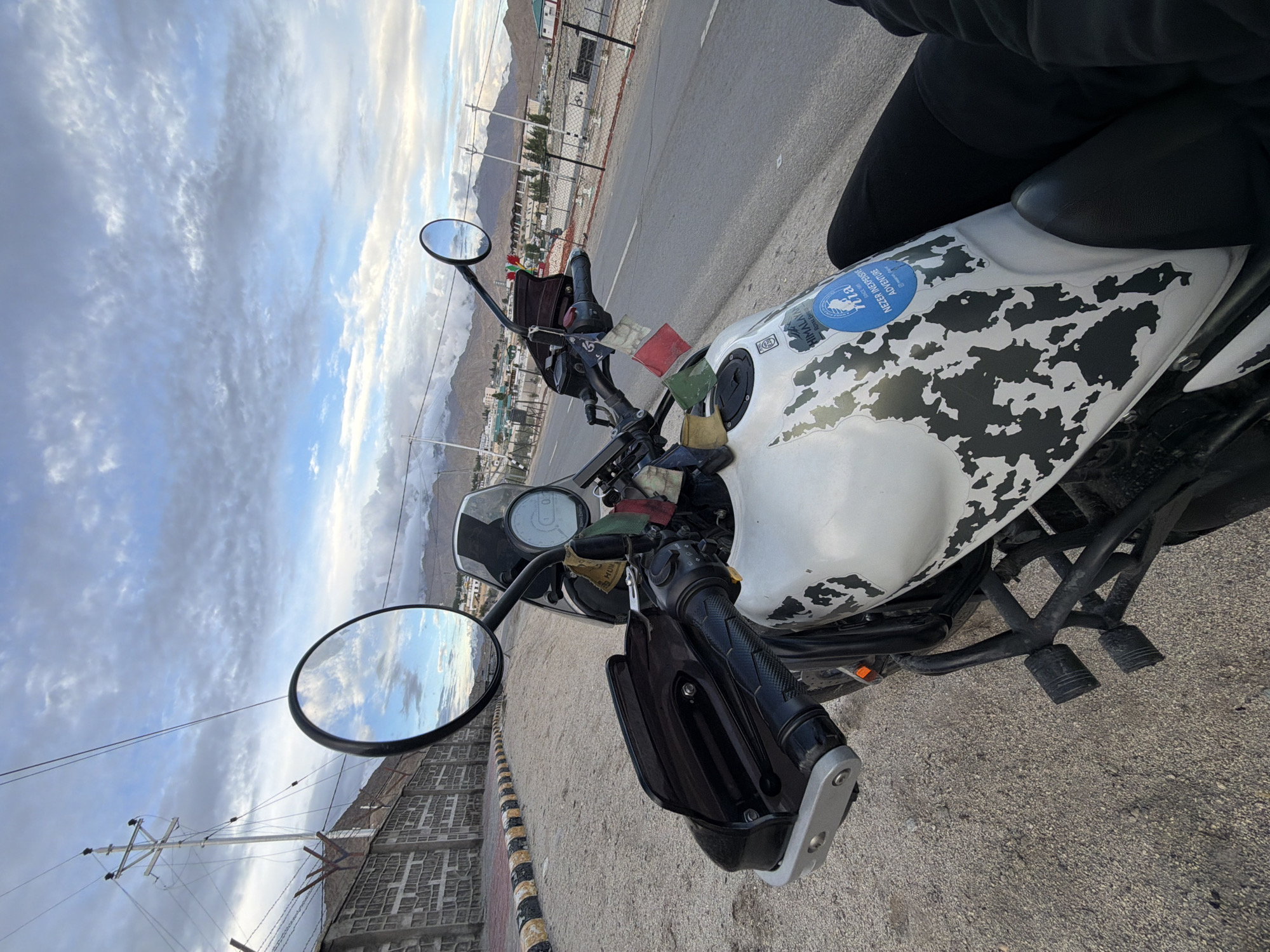 The image shows a white motorcycle with black camouflage-like markings on its fuel tank, parked on the side of a road in Spituk, India. The motorcycle has handguards, two circular mirrors, and several prayer flags attached to its handlebars and engine. A blue circular sticker with text and imagery is also visible on the fuel tank. In the background, a road stretches into the distance, flanked by a low wall with a black and yellow striped border and a chain-link fence. Beyond the fence, there are buildings and mountains under a cloudy sky. A power line pole is visible on the left side of the image. The perspective is from the rider's point of view, with their legs visible in the lower right corner.