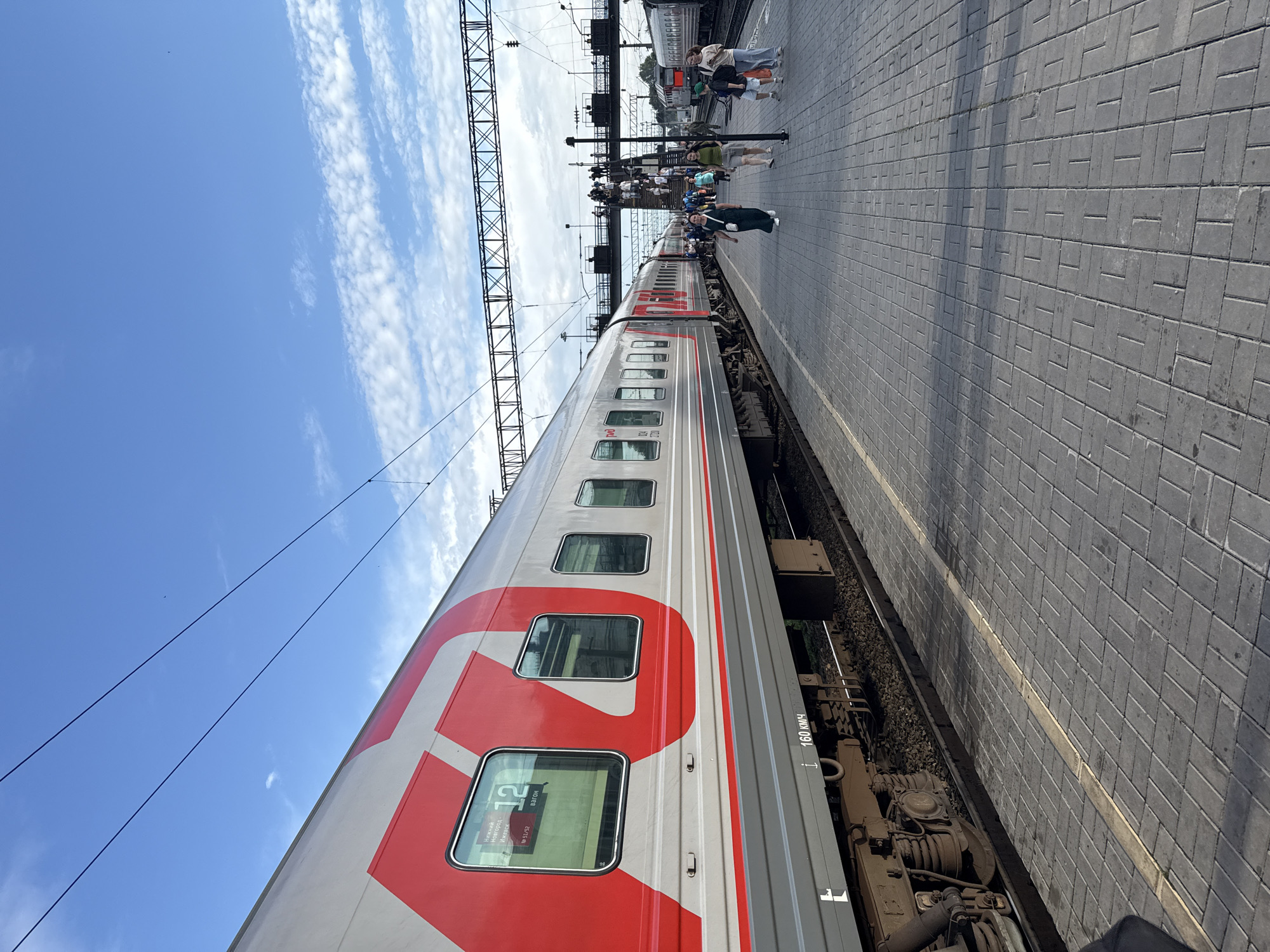 The image captures a train stopped at a station. The train cars are predominantly silver with large red markings. One car prominently displays a large red Cyrillic letter "Р". Several windows on the train are visible, with one window showing the number "12" and the text "Н.Новгород-Москва" (Nizhny Novgorod-Moscow), indicating a route.
On the platform next to the train, several people are present. Some appear to be passengers, possibly waiting to board or disembark, while others might be railway staff. The platform itself is paved with gray interlocking bricks. Overhead, electrical wires for the train are visible, supported by metal structures.
The sky is a bright blue with scattered white clouds, suggesting it is daytime with fair weather. The sun is casting shadows on the platform. The overall scene depicts a typical moment at a train station in Izhevsk, Russia, during a clear day. The presence of passengers and the train suggest an active railway environment. The visible text on the train provides specific information about its destination.