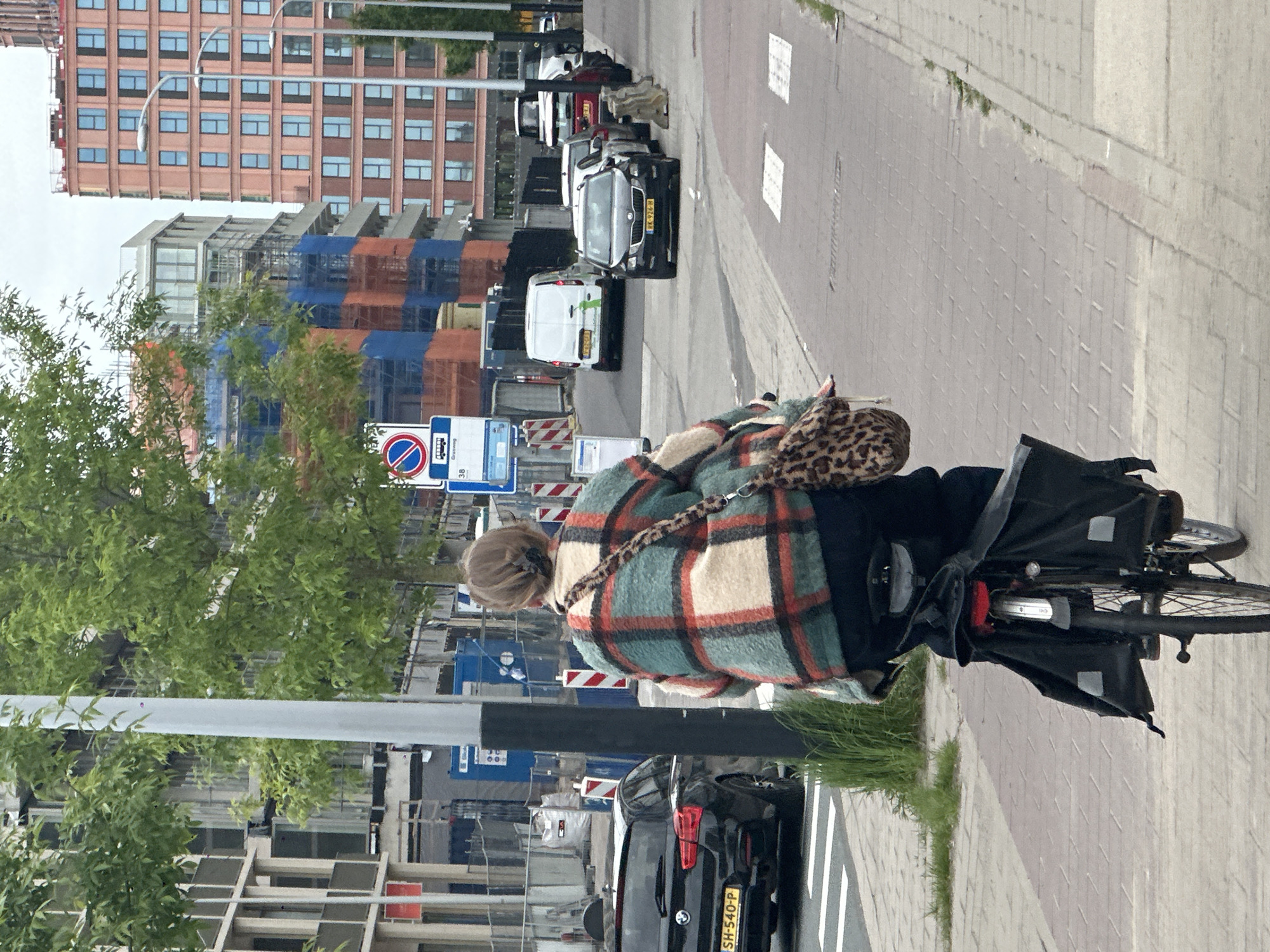 The image captures a street scene in Amsterdam, Netherlands. In the foreground, a woman rides a bicycle along a brick-paved sidewalk. She wears a plaid jacket and a leopard-print bag. The bicycle has black panniers attached to the rear. Behind her, a row of parked cars lines the street. Among them, a black BMW is visible with a license plate reading "SH-540-P." Across the street, construction is underway, with buildings wrapped in blue and orange material. A sign restricting truck parking is visible. Taller brick buildings rise in the background under a cloudy sky. A streetlight is visible to the woman's left.