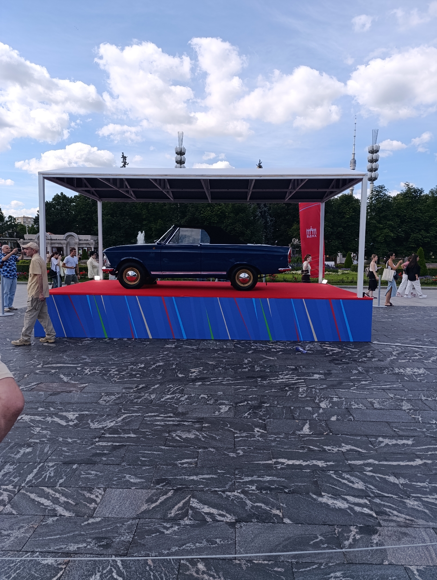 An outdoor event is taking place under a bright, cloudy sky. A dark blue convertible car is displayed on a raised platform covered with a red carpet. The platform is adorned with blue panels featuring red, white, and green diagonal stripes. To the left of the platform, a man in a light tan shirt and pants walks past, while another man in a blue floral shirt takes a photo. Several other individuals are visible in the background, some walking and others observing the display. Behind the platform, trees form a dense green backdrop, and a red banner with white Cyrillic text is visible. The ground is paved with large, dark gray, and white marbled tiles. The scene appears to be in an urban park or plaza.