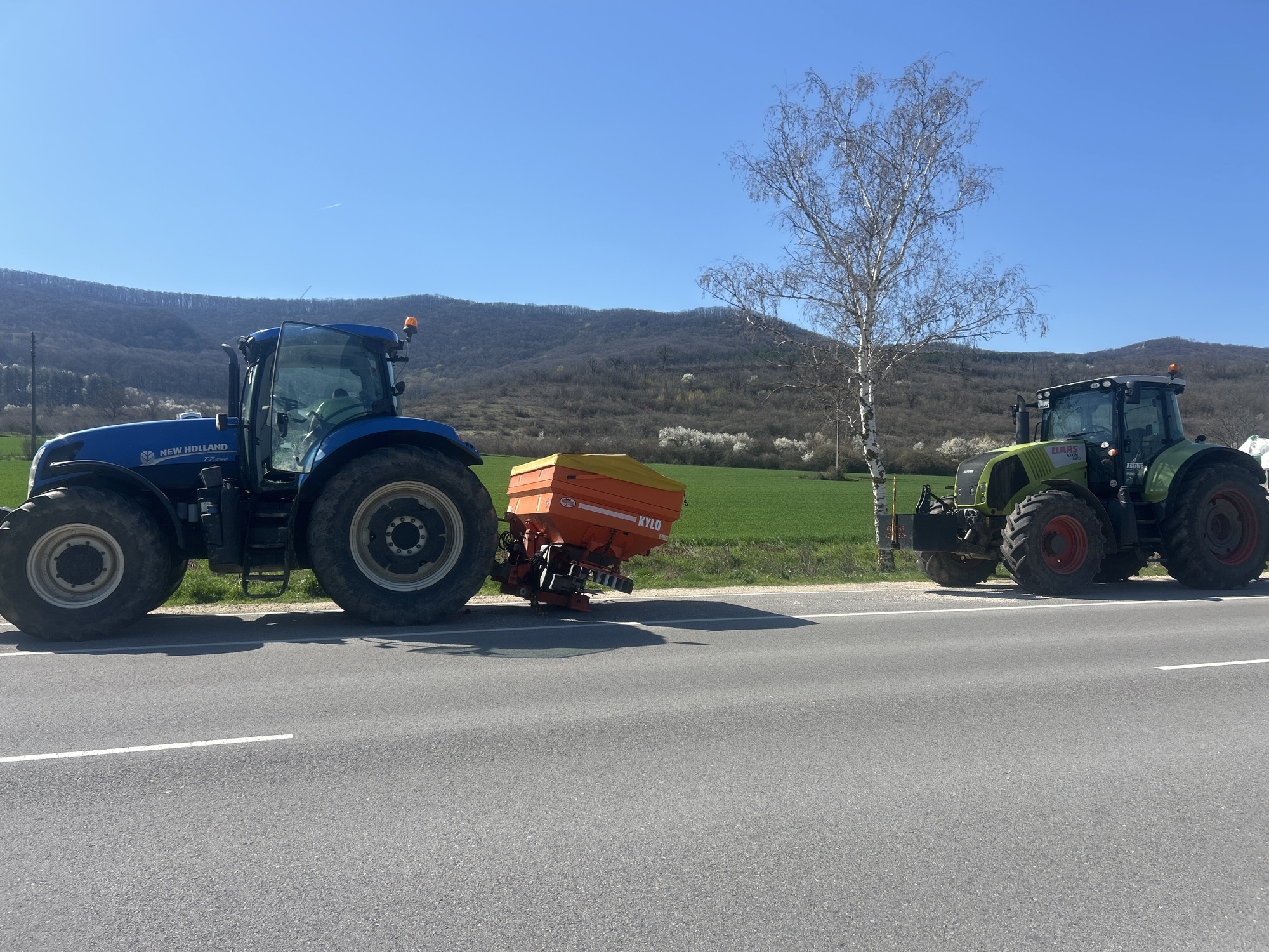 The image shows a rural roadside scene with two tractors parked on the asphalt. The road has a dividing white line. The tractor on the left is a blue New Holland, and it has a yellow and orange spreader attached to the back. The tractor on the right is green and bears the brand "CLAAS AXION". In the background, there is a green field, a bare tree, and hills. The sky is blue and clear. The location is likely near the city of Gradishte, Bulgaria, based on the prompt.