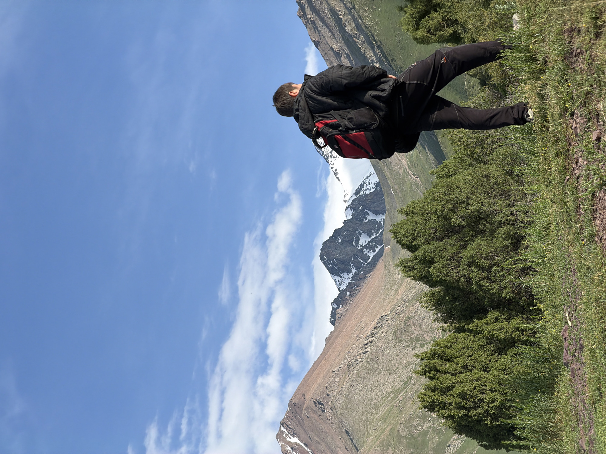 A person with a black jacket and a red backpack stands on a grassy slope overlooking a mountain range. The person is facing away from the camera, looking out at the vast landscape. The mountains are rugged and appear to be covered in snow on their peaks, contrasting with the green valleys and slopes below. The sky is a clear blue with a few wispy clouds. The terrain suggests a high-altitude, mountainous environment, typical of a hiking or trekking location. The lighting indicates it's daytime, likely mid-morning or afternoon, with bright sunlight illuminating the scene. The overall mood is one of awe and contemplation of nature's grandeur. There is no visible text in the image. The location context of Kashka Suu, Kyrgyzstan, further supports the interpretation of a remote and scenic mountainous region.
