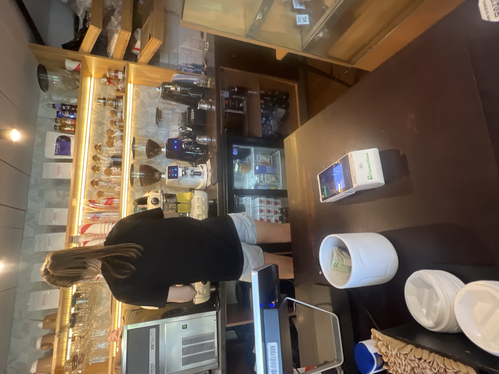 In a coffee shop in Fontanka, Ukraine, a young woman with her hair in a ponytail stands behind a counter, facing away from the camera. She is wearing a black t-shirt and light-colored shorts. In front of her, on the counter, are various coffee-making appliances, including grinders and a coffee machine. To her left, an ice machine is visible.
The counter is cluttered with items typically found in a cafe: a payment terminal, a white ceramic cup filled with cash, stacks of white disposable lids, a tray of what appears to be biscuits or crackers, and a small blue container. Behind the woman, shelves are stocked with bagged coffee, glassware, and bottles. A refrigerator unit is visible beneath the counter, containing cartons of milk and bottled drinks. To the right of the scene, a display case with shelves is partially visible, with price tags indicating "65" and "75". The overall environment suggests a busy but organized coffee shop setting.