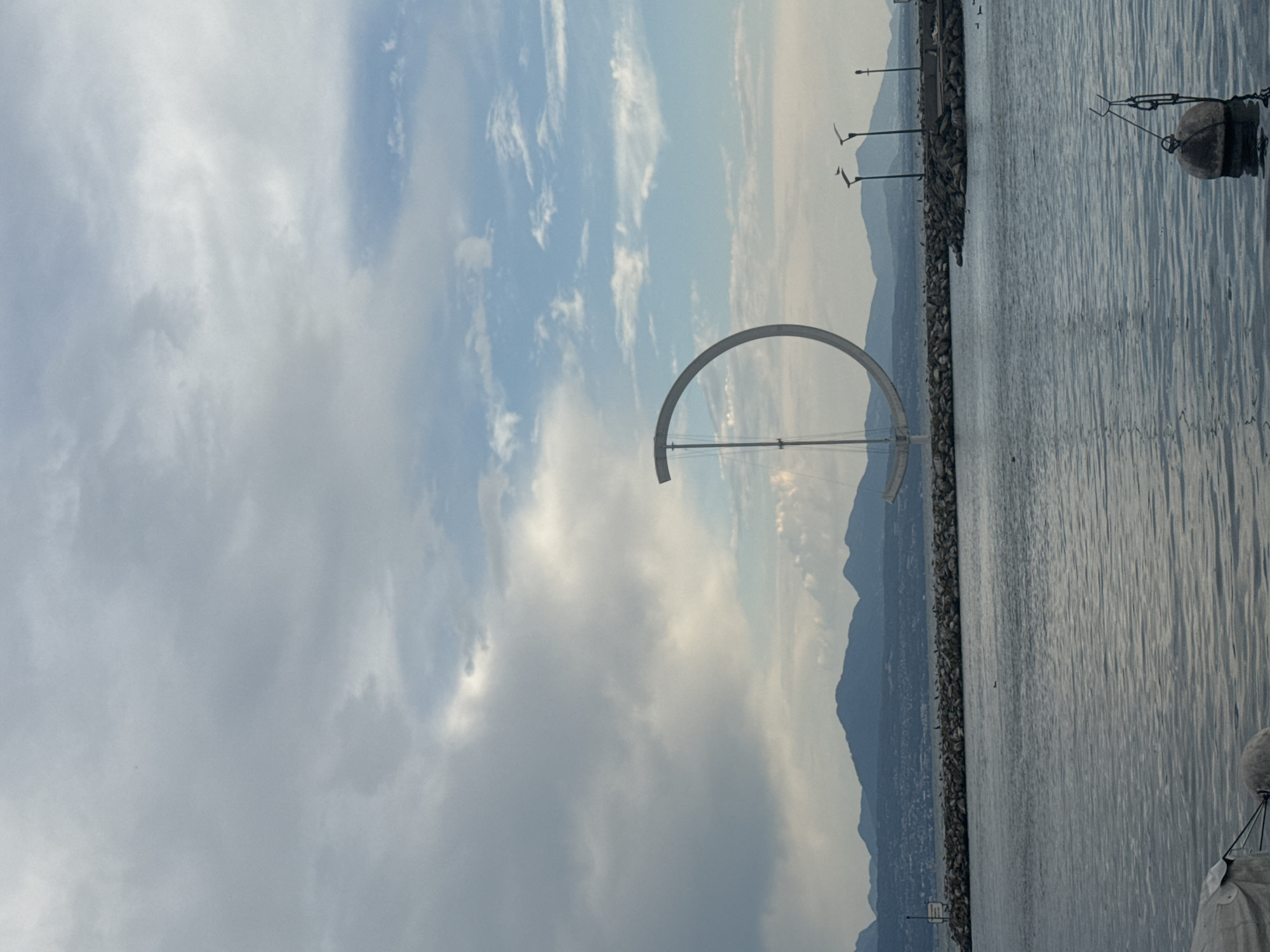 The image depicts a large, circular sculpture with a vertical support pole, resembling a stylized euro symbol, situated in the water near the shoreline of Lake Geneva in Lausanne, Switzerland. The sculpture is positioned in the foreground, slightly to the right of the center of the frame. The water is calm, with ripples reflecting the overcast sky.
In the background, a man-made breakwater made of large rocks lines the edge of the lake. Beyond the breakwater, a city skyline is visible, nestled against a range of distant, hazy mountains. Several streetlights with a distinctive, modern design are spaced along the breakwater. The sky is predominantly gray and cloudy, with patches of blue sky visible, indicating a partly cloudy day. The overall atmosphere is serene and slightly moody due to the weather conditions. There are no people clearly visible in the image.