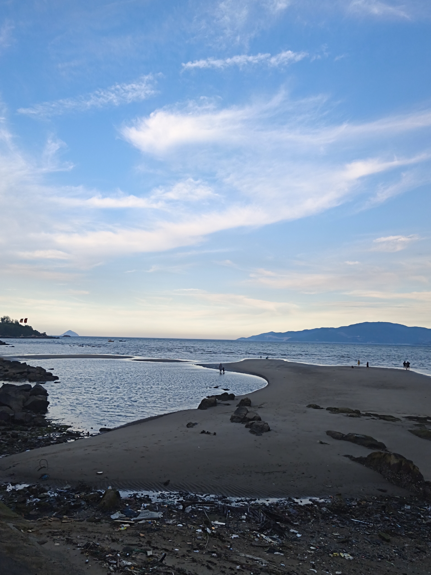 This image depicts a coastal scene in the Bac Nha Trang Ward, Vietnam. The foreground is dominated by a dark, sandy beach littered with visible debris, including plastic waste and wood fragments. A small body of calm water, possibly a tidal pool or inlet, separates this cluttered area from a larger expanse of the sea.
Further out, a narrow sandbar curves into the water, with several people walking along its crest. The sea stretches to the horizon, meeting a distant mountainous coastline under a pale blue sky filled with wispy white clouds. Two small boats are visible on the water. In the middle ground, on a rocky outcrop to the left, two red flags are planted. The overall impression is a mix of natural beauty and human-generated pollution.