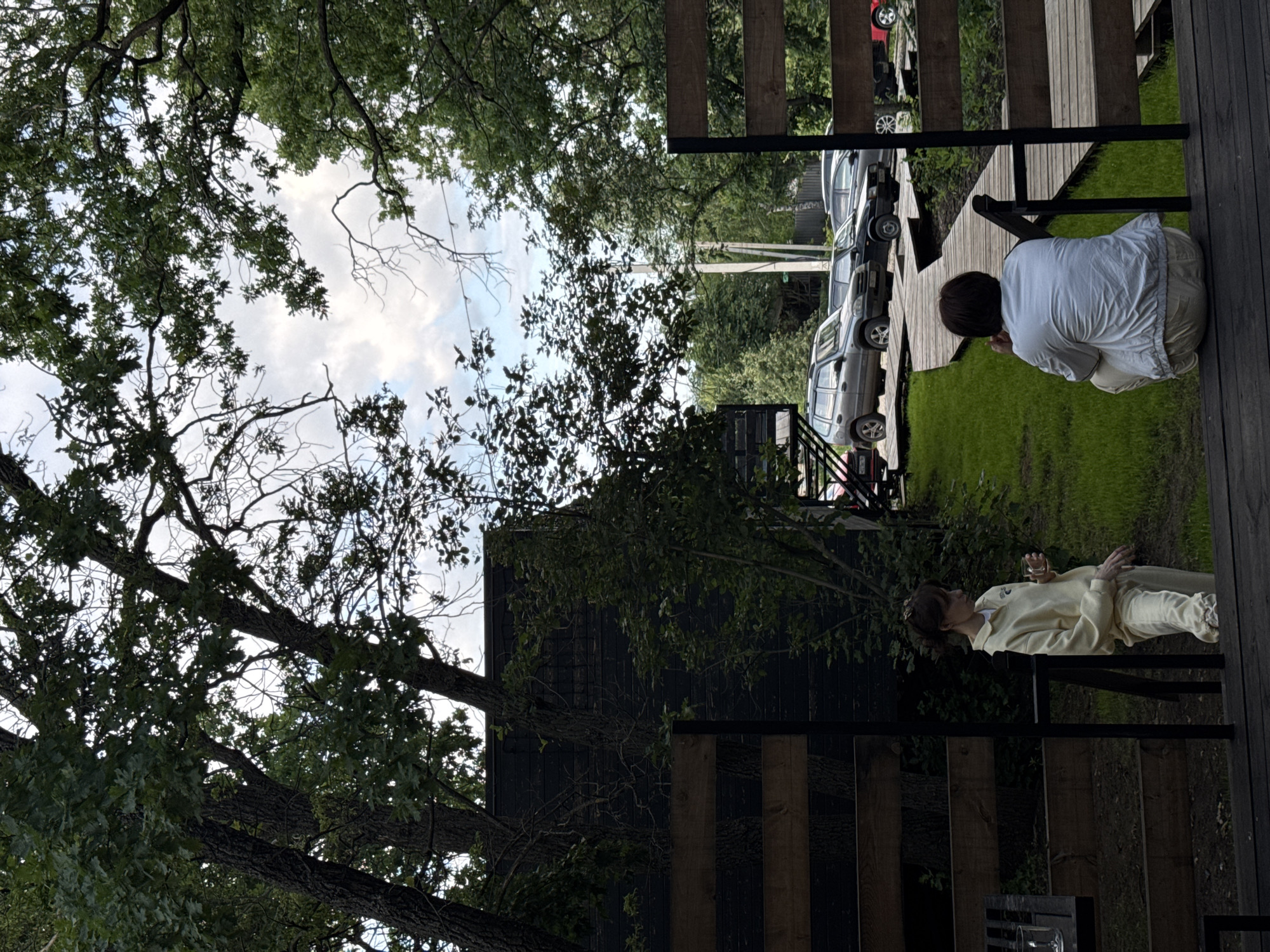 The image depicts an outdoor scene in Ust-Kurdyum, Russia. A woman wearing a light yellow tracksuit is standing near a wooden fence and holding a glass. Further to the right, another person is crouched on a wooden platform, facing away from the camera. A winding wooden walkway is visible on a grassy area in the background, where several cars are parked. The environment is characterized by mature trees and overcast skies.