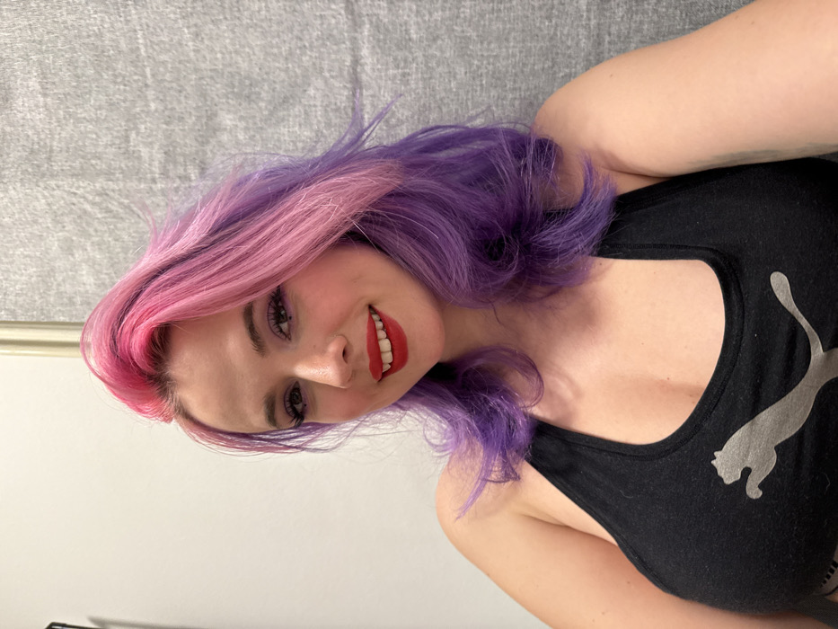 A woman with pink and purple hair smiles at the camera, showcasing red lipstick and a nose piercing. She is wearing a black tank top with a large silver Puma logo. The background includes a gray textured wall and a glimpse of a white door frame. The photo is rotated, giving a tilted perspective.