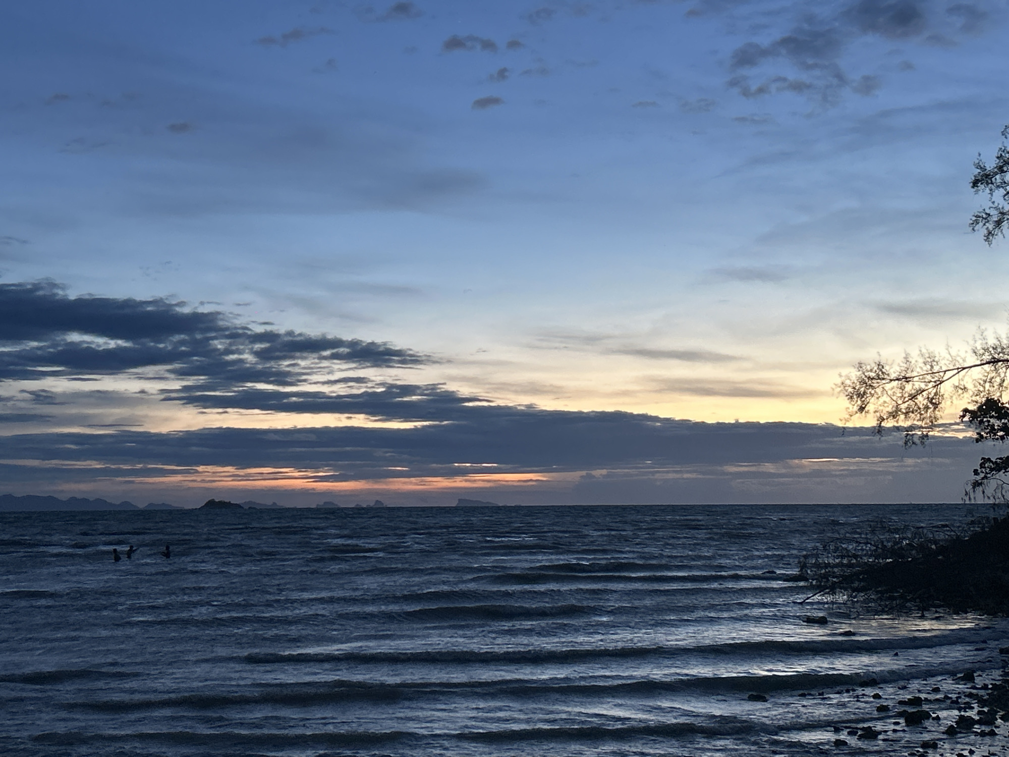 The image depicts a seascape at dusk, likely in Baan Sri Thanu, Thailand, given the prompt. The sky is a gradient of blues and greys, with clouds scattered across the horizon, some tinged with yellow from the setting sun. The sea is choppy with small waves. In the distance, silhouettes of small islands are visible. In the water, closer to the shore, there are three figures of people, presumably swimming or wading. The right side of the frame features dark foliage and the branches of a tree, obscuring the view of the land. The foreground shows the beach with small rocks.