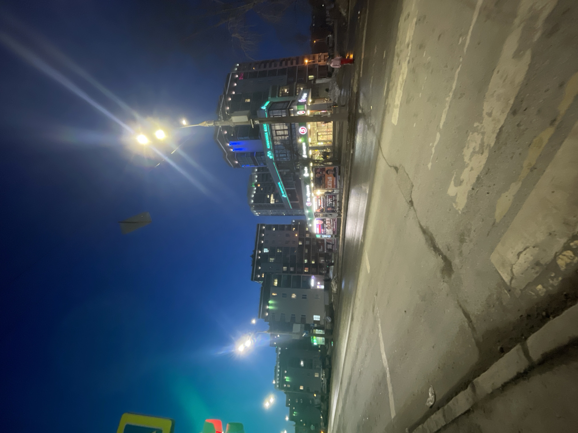 The image shows a street scene at night in what appears to be Izhevsk, Russia. A multi-story building complex dominates the background with illuminated windows and commercial signage in both Russian and English. Bright streetlights illuminate the scene, casting flares into the dark blue sky where a paper airplane is visible. A wide, cracked sidewalk occupies the foreground with faded pedestrian crosswalk markings. The pavement appears wet, reflecting the ambient light. At the top right, a person in a pink jacket walks on the sidewalk. Traffic signs are seen at the bottom left.