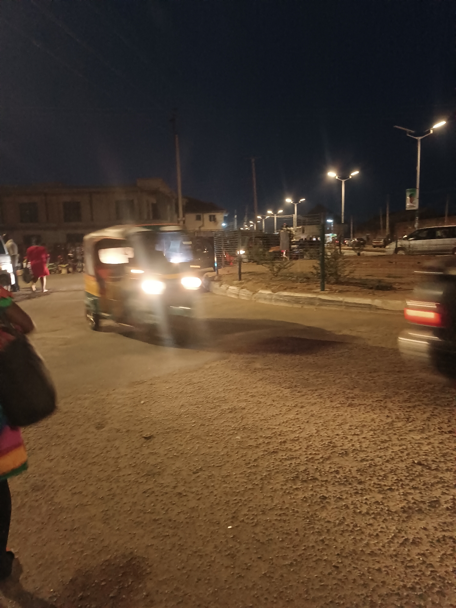 The image depicts a street scene in Bukuru, Nigeria, at night. An auto rickshaw, or "keke," with headlights on, is the central subject, moving on a paved road. A person wearing a red dress is walking on the left side of the road. Other vehicles are visible in the background. The scene is lit by streetlights and the headlights of the rickshaw. A fence, landscaping and a small building can be seen on the right.