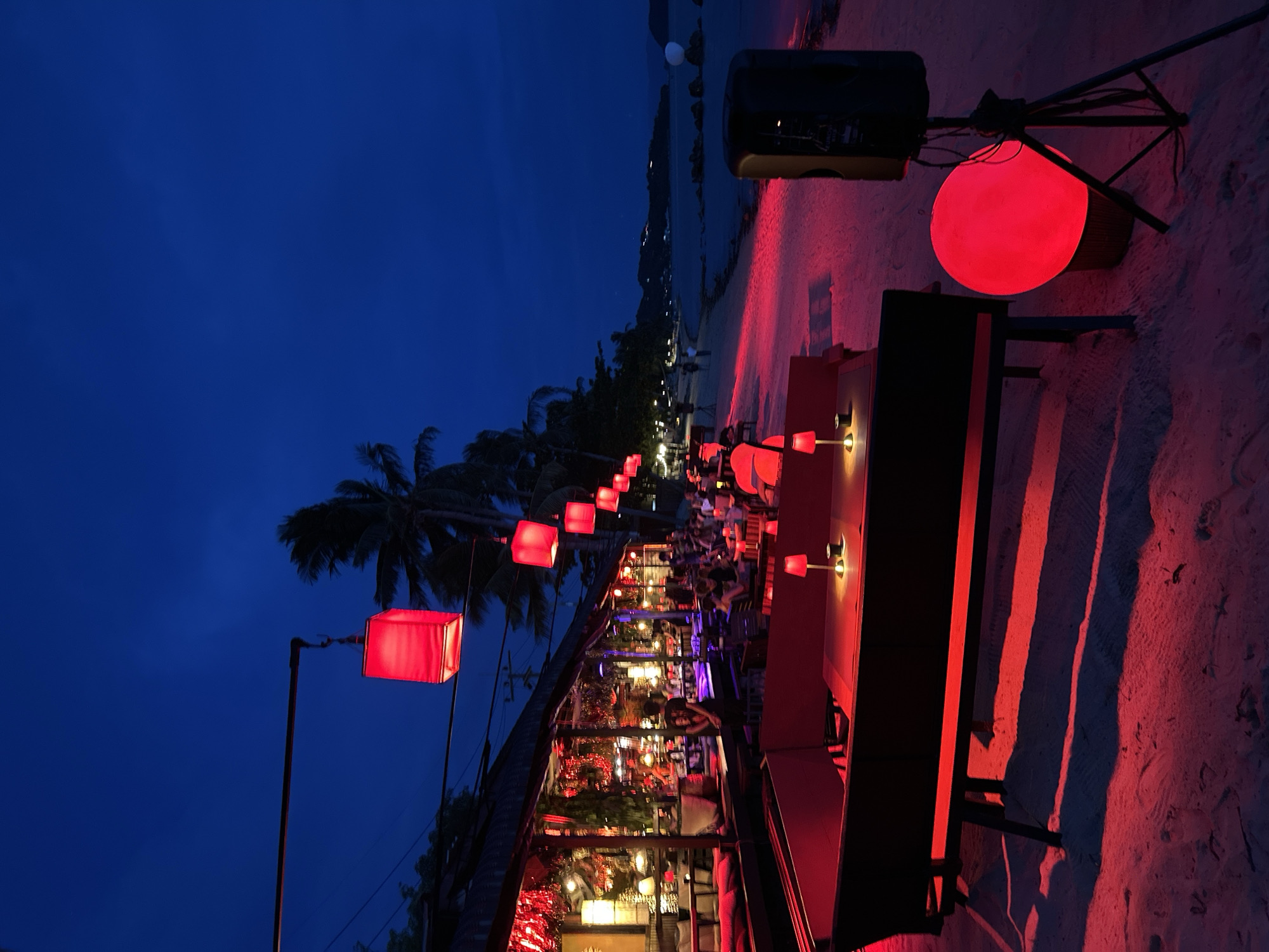 The image depicts a beachside restaurant at dusk or night on Ko Pha-ngan, Thailand. The scene is illuminated by an abundance of red lights, creating a vibrant and festive atmosphere. Row of square red lanterns hang suspended from poles, lining the walkway leading to the restaurant, while a large, spherical red light sits near a speaker and a dark, rectangular structure on the sandy beach.
The restaurant itself appears to be a covered open-air structure, with tables and chairs set up. People are seated at the tables, suggesting a dining or social gathering is in progress. The ambiance is warm and inviting, with the red lighting casting long shadows across the sand. Palm trees are visible in the background, further indicating a tropical location. The sky is a deep, clear blue, characteristic of twilight or nighttime. The overall impression is one of a lively evening at a popular beach establishment.
